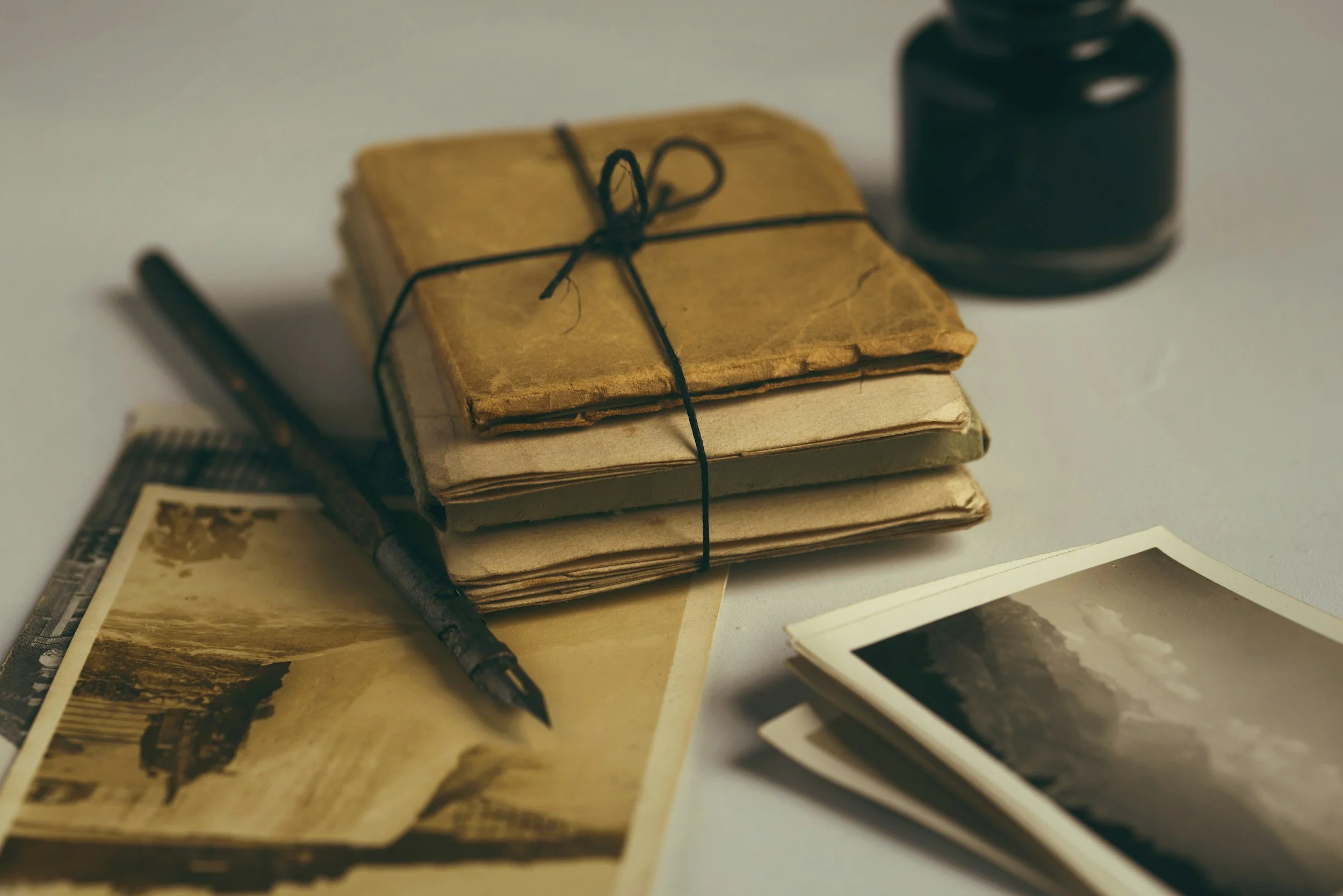 Stack of old, worn books tied with black string, a black fountain pen, and vintage photographs spread on a white surface.