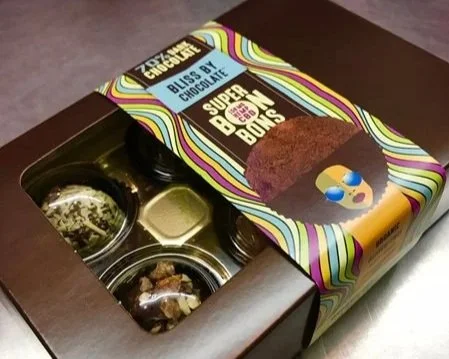 Open box of assorted chocolates and chocolates with colorful packaging labeled 'Chocolatier Collection Blis by Chocolate' and 'Super Buns'.