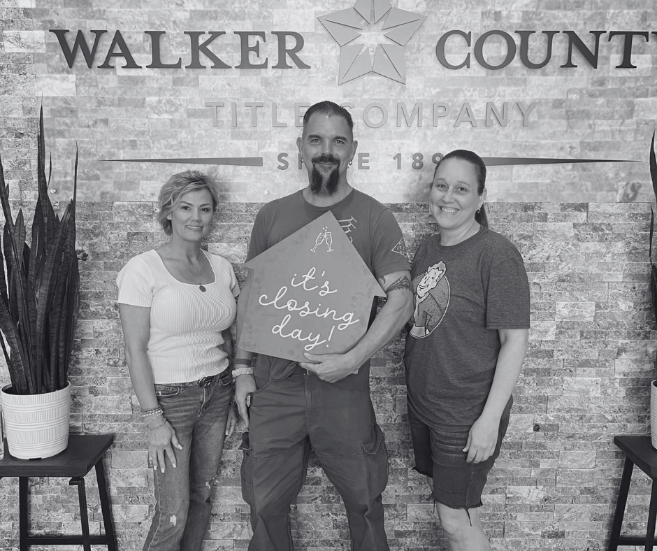 Three people standing in front of a brick wall with the logo of Walker County Title Company. The man in the middle is holding a sign that says 'It's closing day!' and is smiling, flanked by two women, all smiling for the camera.