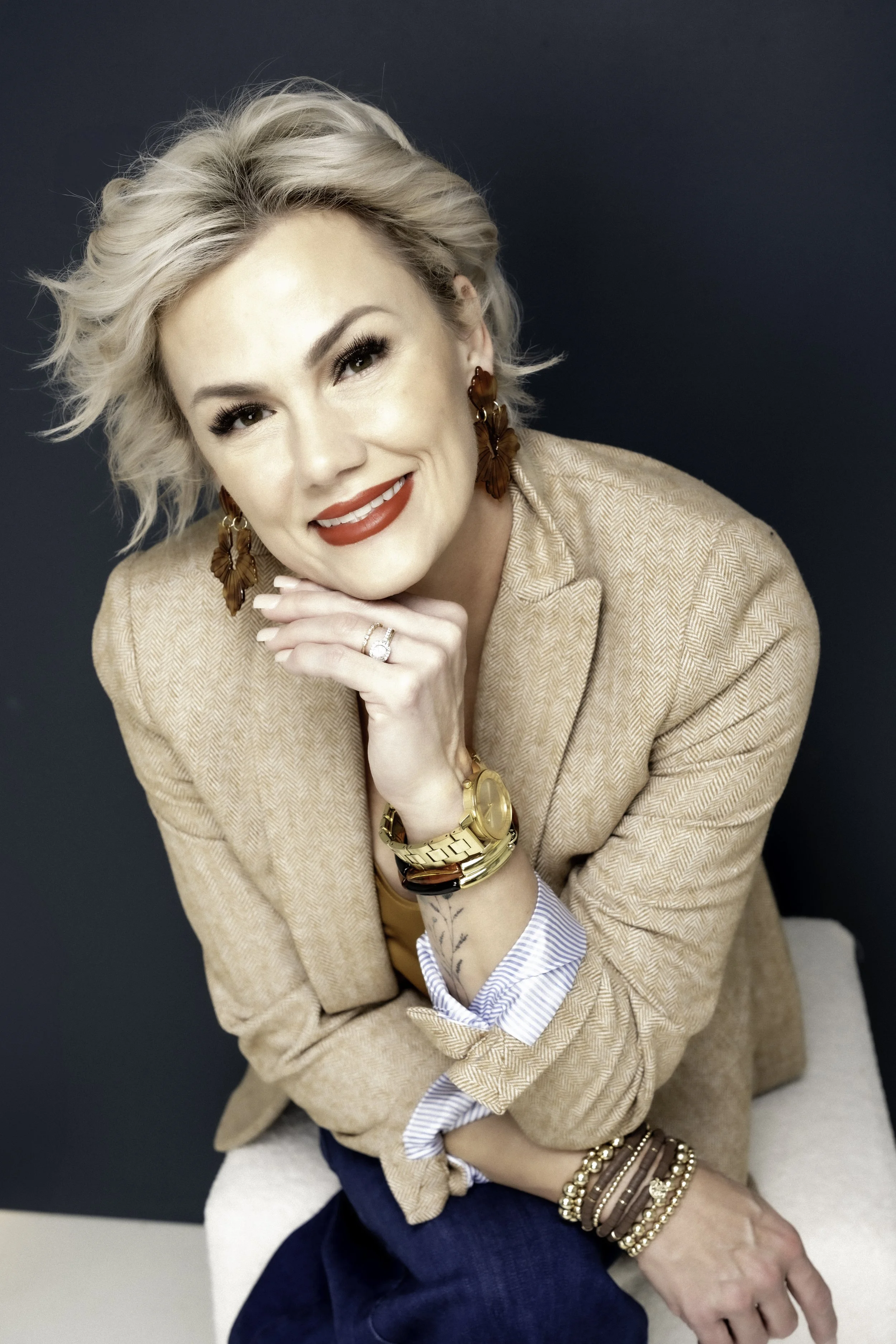 A woman with blonde, wavy hair and red lipstick smiling, wearing a tan blazer, gold jewelry, and dark blue jeans, sitting against a navy background.