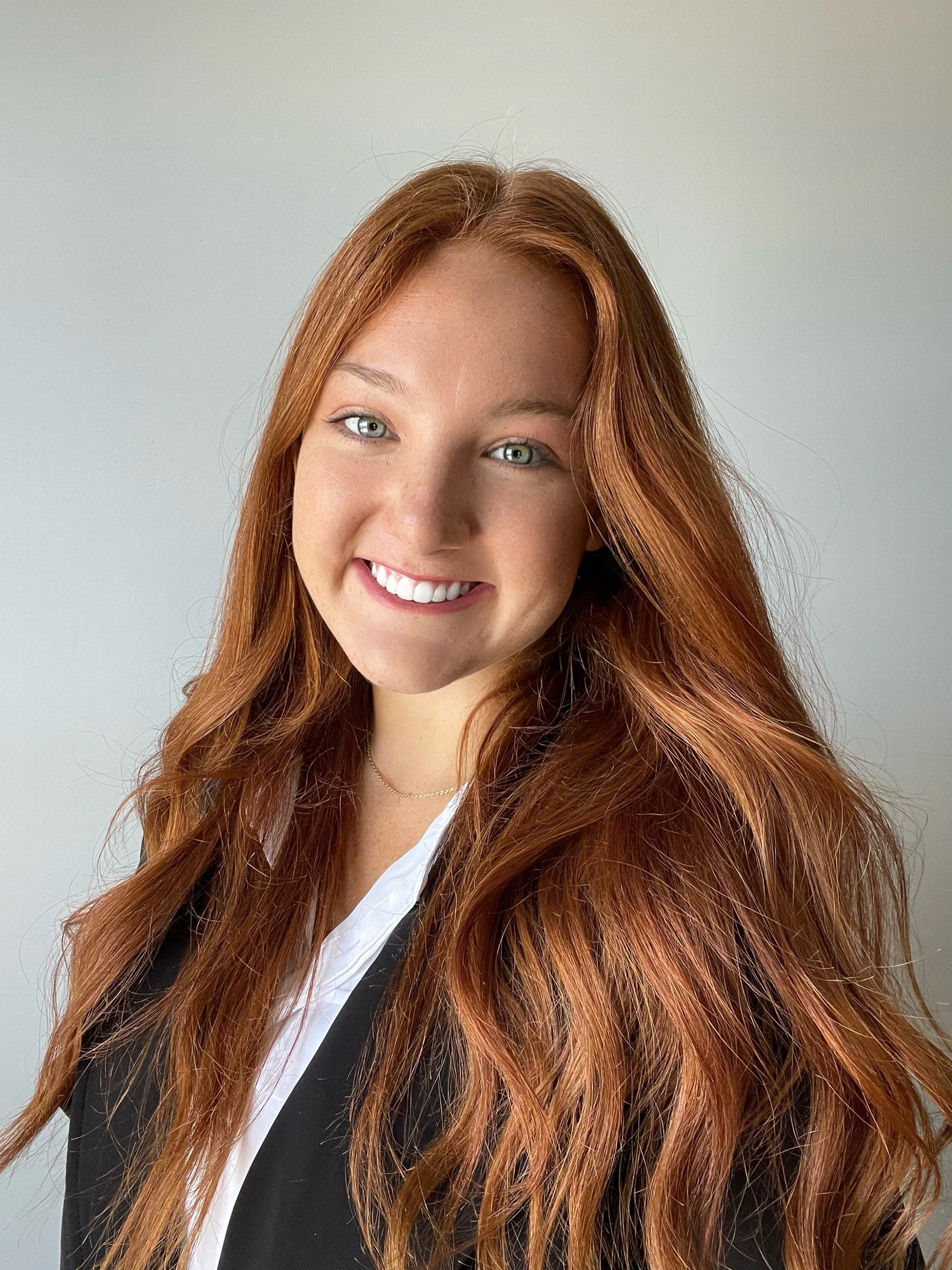 A young woman with long, wavy red hair, light skin, and bright blue eyes, smiling showing her teeth, wearing a black blazer over a white shirt, standing against a light gray background.