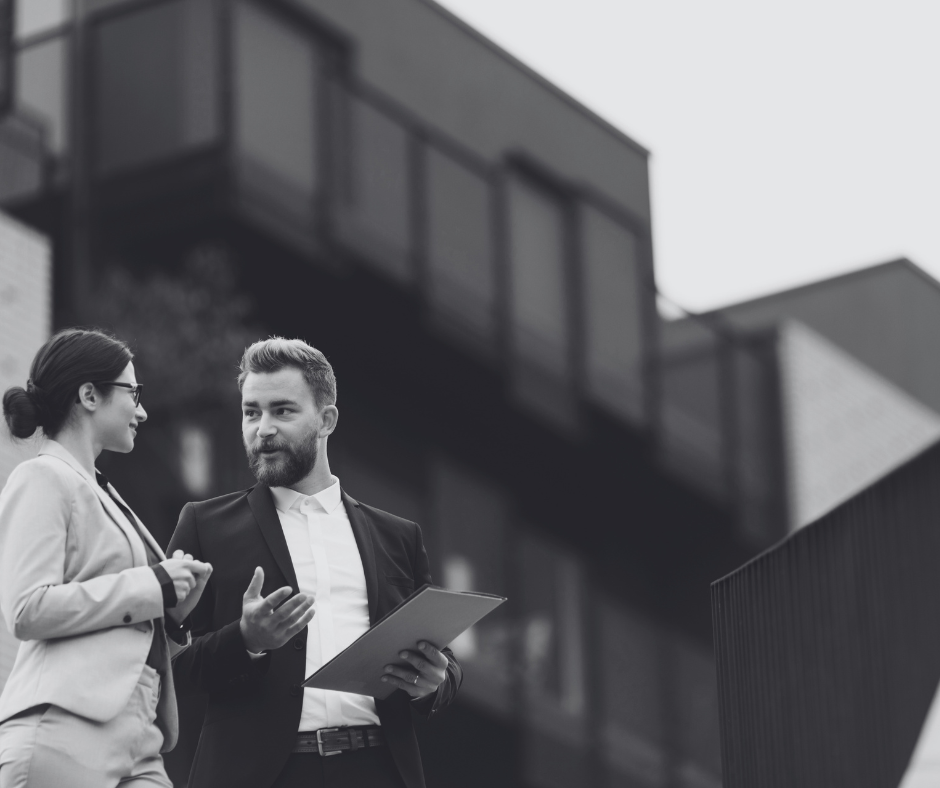 Two business professionals, a woman wearing glasses and a man with a beard, conversing outdoors with a modern building in the background.