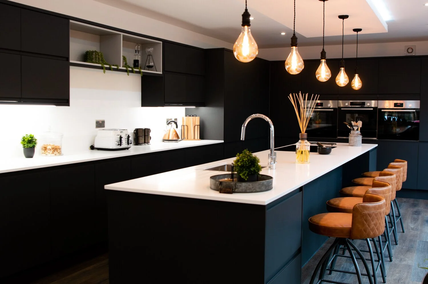 Modern kitchen with black cabinetry, white countertop island with brown leather bar stools, pendant lights, and built-in appliances.