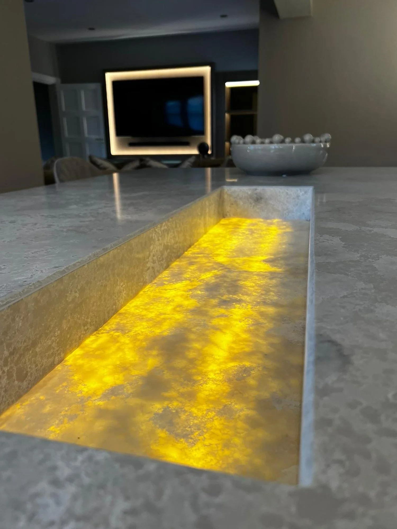 Close-up of a modern kitchen island with a yellow illuminated strip underneath, a large bowl with decorative spheres, and a professional-looking television in a dark living room in the background.