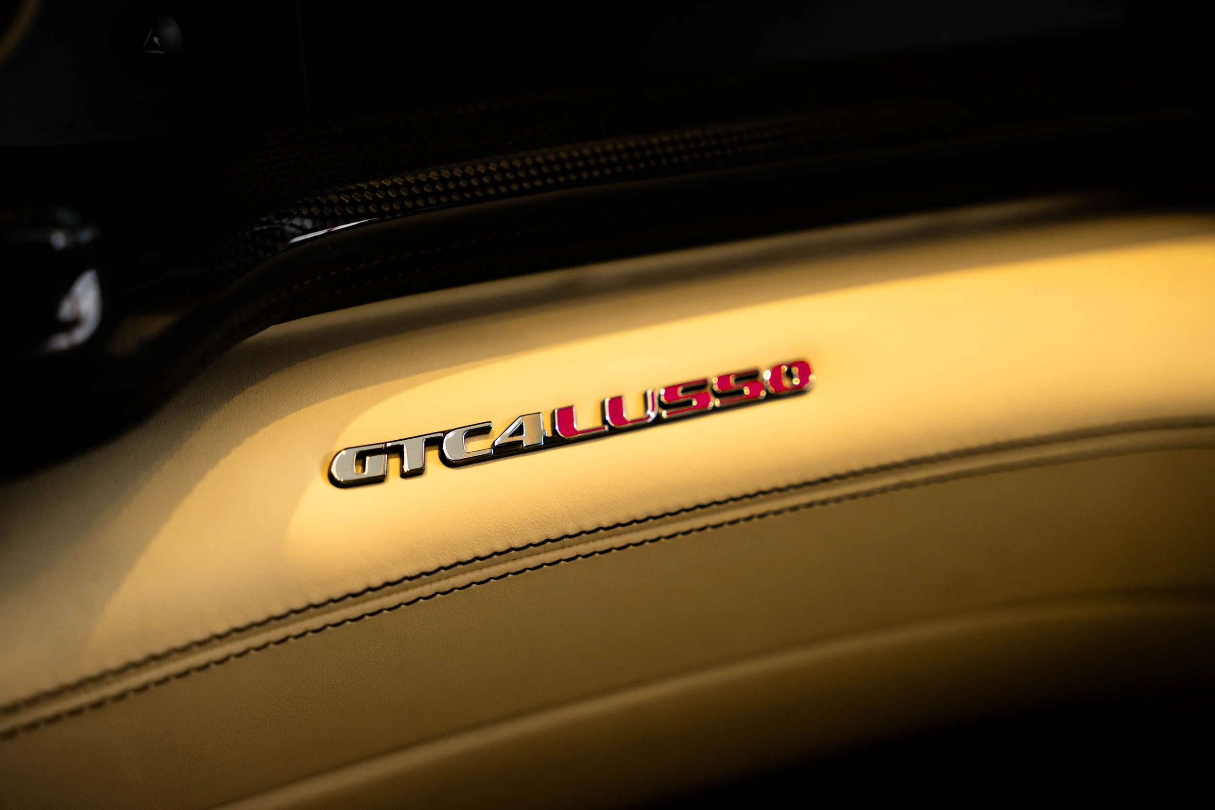 Close-up of a leather surface with a metallic emblem spelling 'GT4LUSSO' and a carbon fibre dashboard above it.