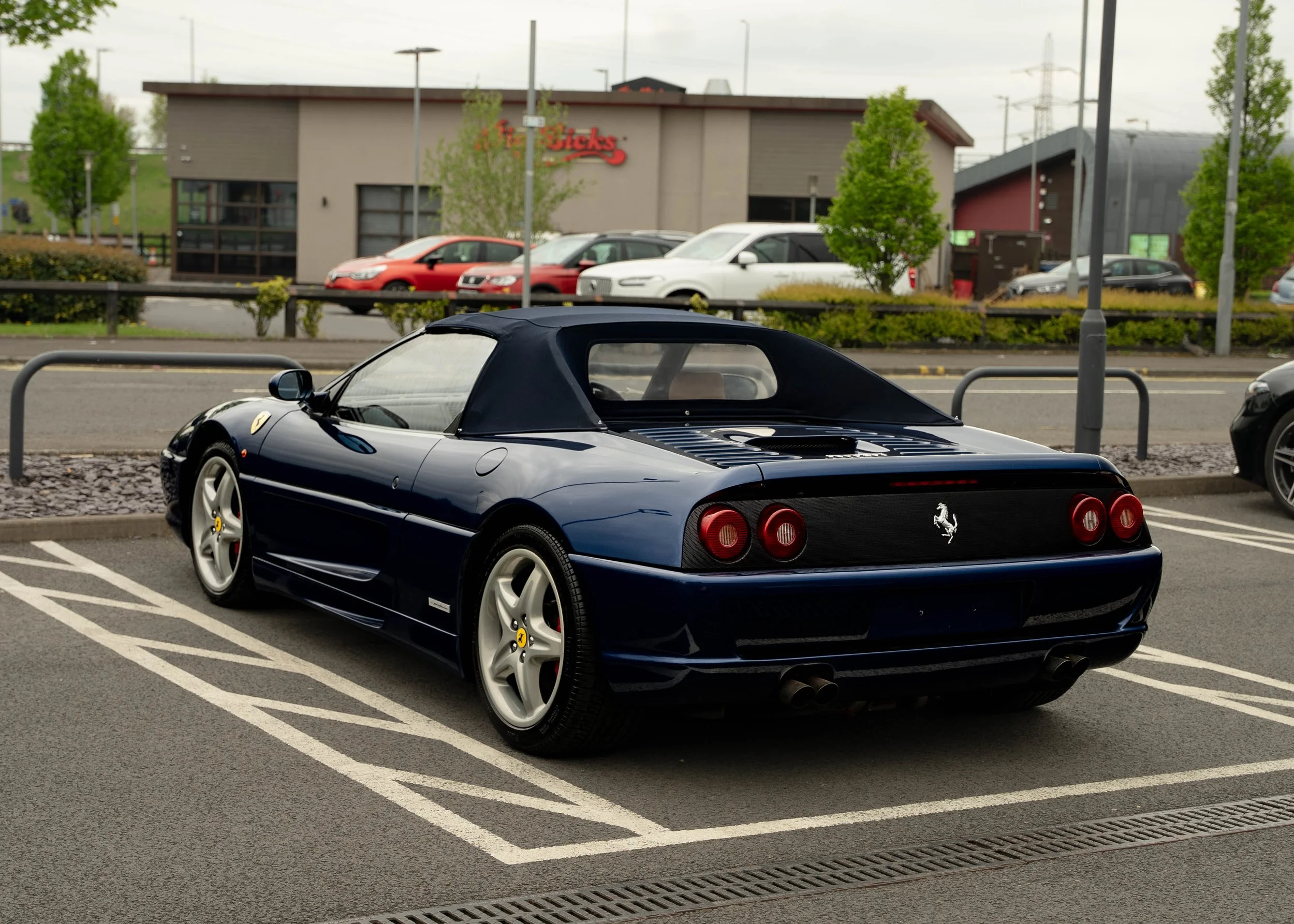 A black Ferrari sports car with a convertible top parked in a parking lot. The background shows a shopping center with parked cars, trees, and a building with a sign that reads "Red Sticks." The sky is overcast.