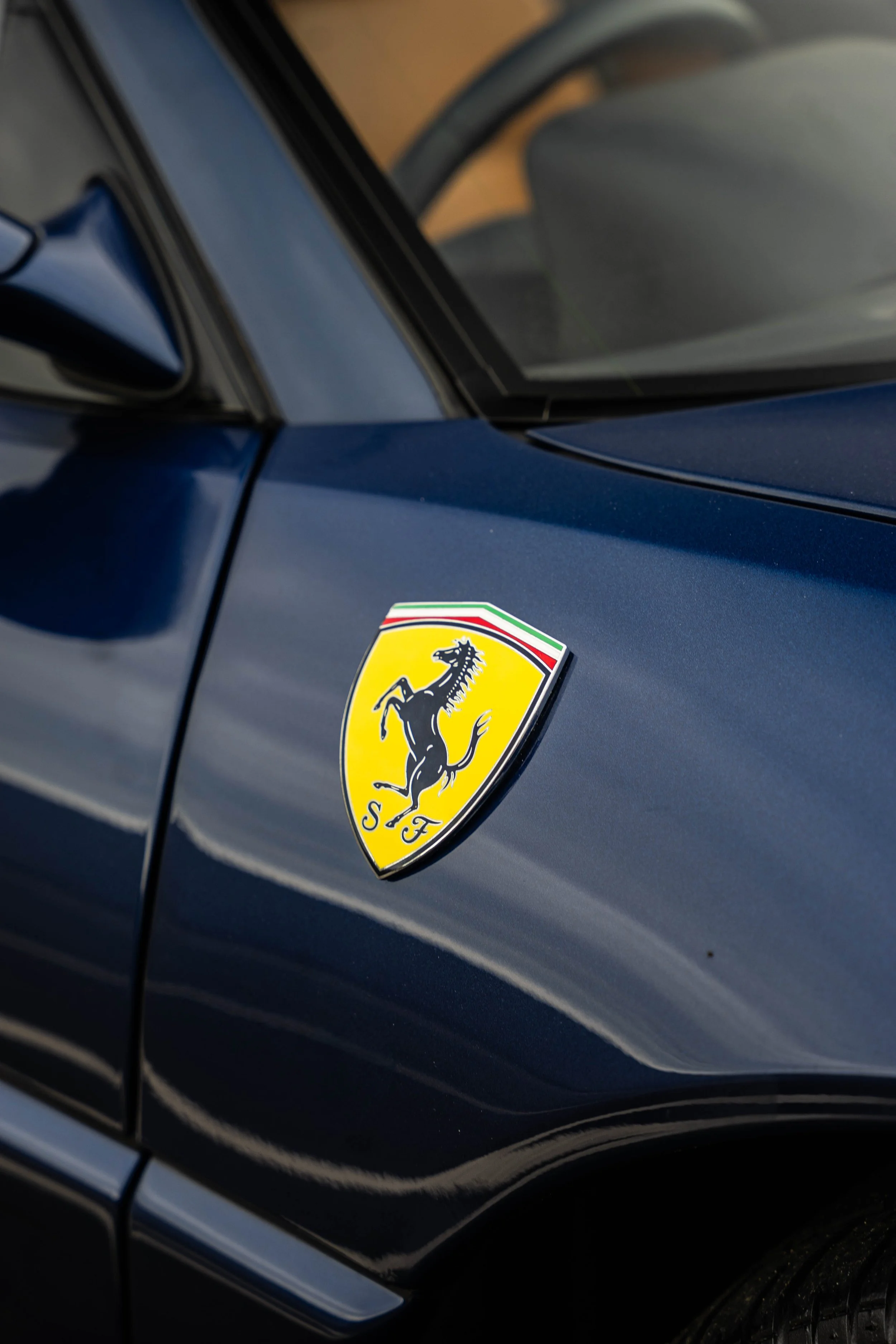 Close-up of a dark-colored Ferrari car with a yellow shield-shaped badge featuring a black prancing horse and the letters S F.
