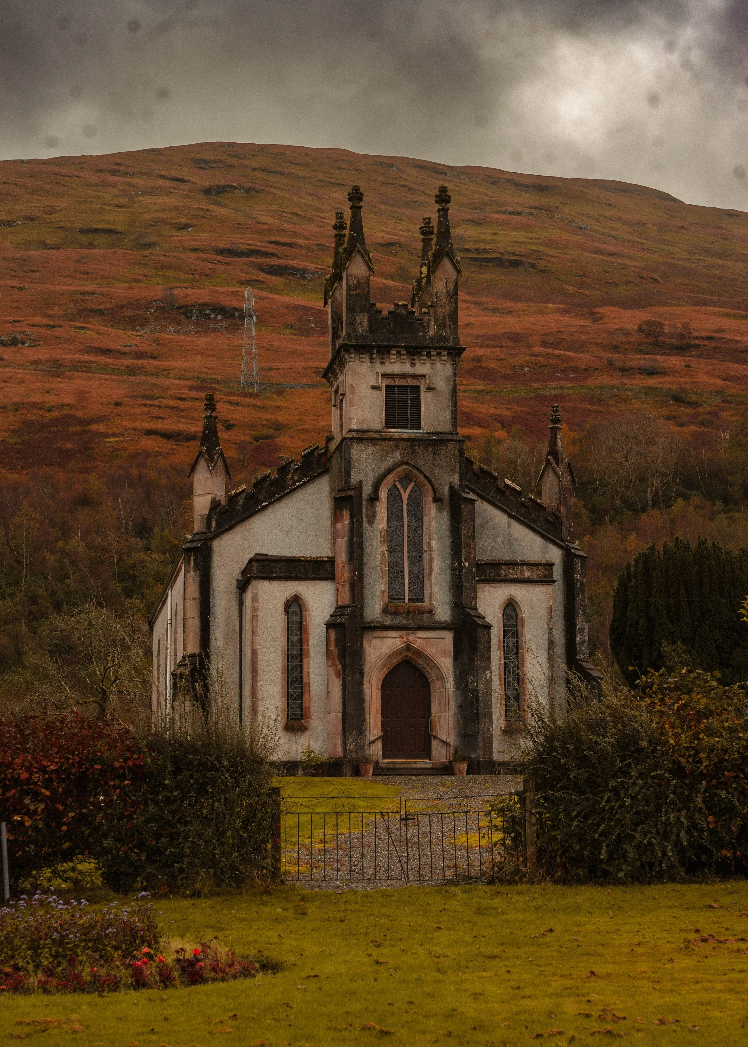 An old church with gothic architecture, pointed windows, and spires, surrounded by a garden, with a mountain covered in reddish-brown vegetation in the background under a cloudy sky.