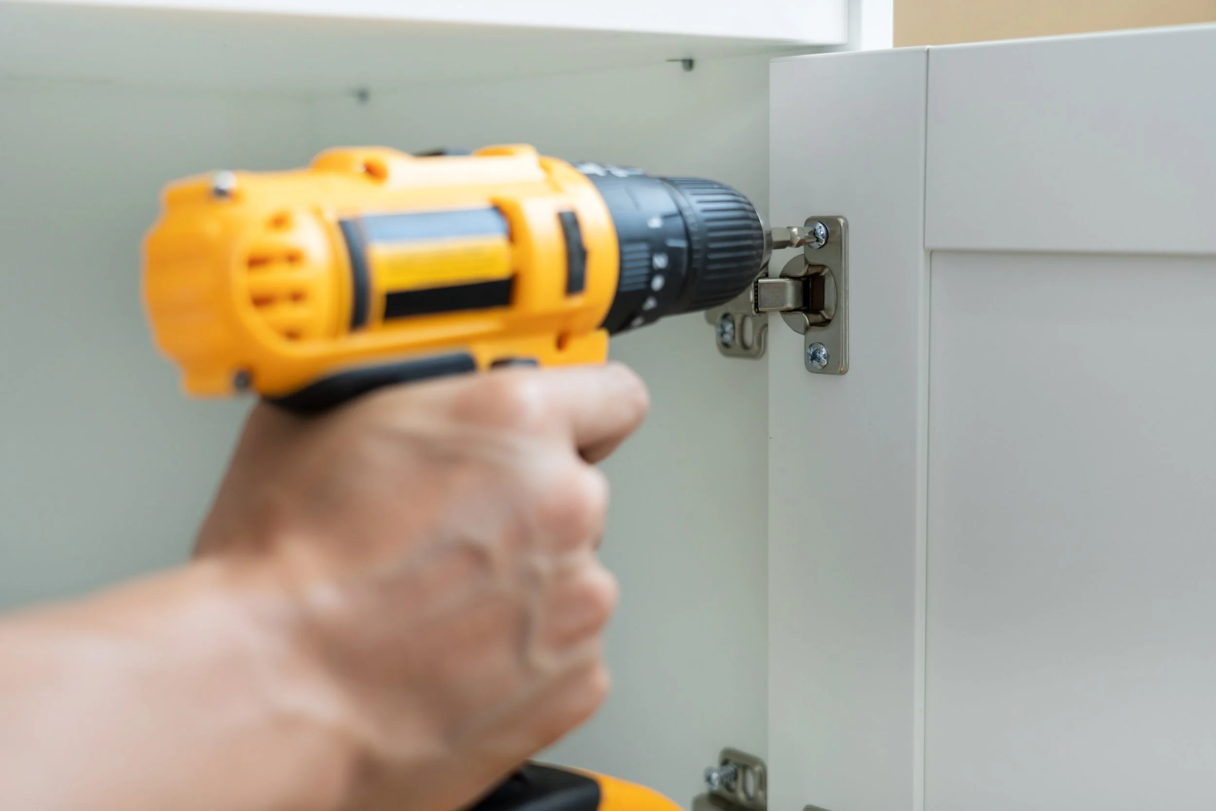 A person using a yellow cordless drill to attach a silver latch to a white cabinet door.