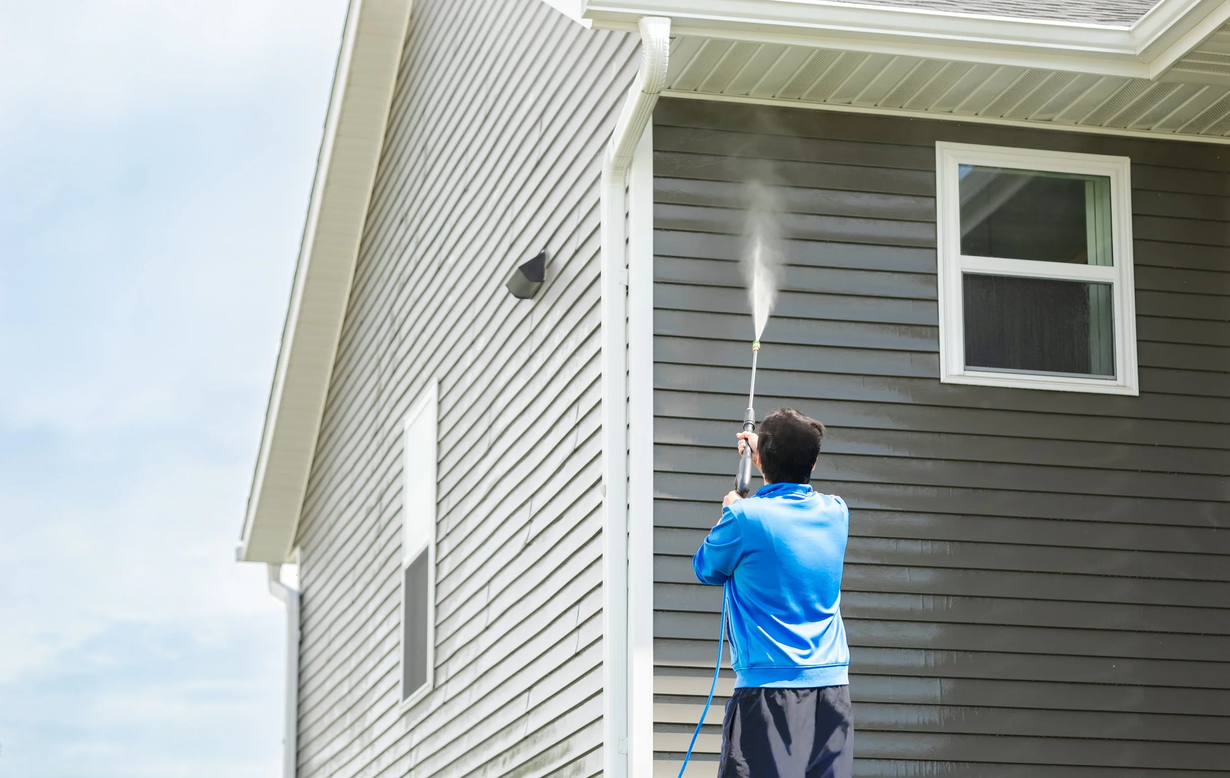 Man in blue jacket pressure washing the side of a house with gray siding