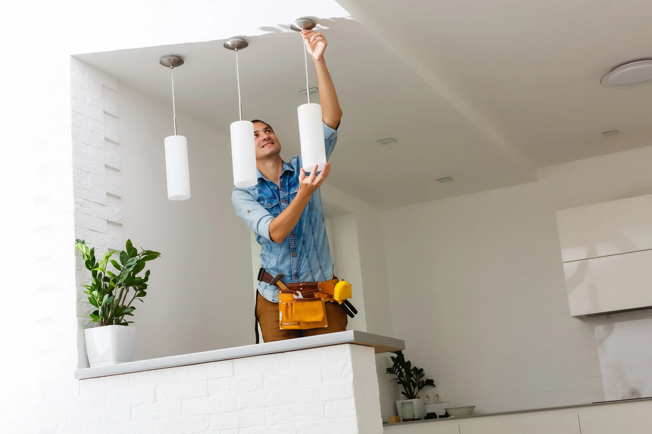 A man in a denim shirt and brown pants installing a modern white pendant light fixture in a home interior. He is using a screwdriver and wearing a tool belt with various tools. The room has white walls, ceiling, and minimal decor, including a potted plant and some small objects on a shelf.