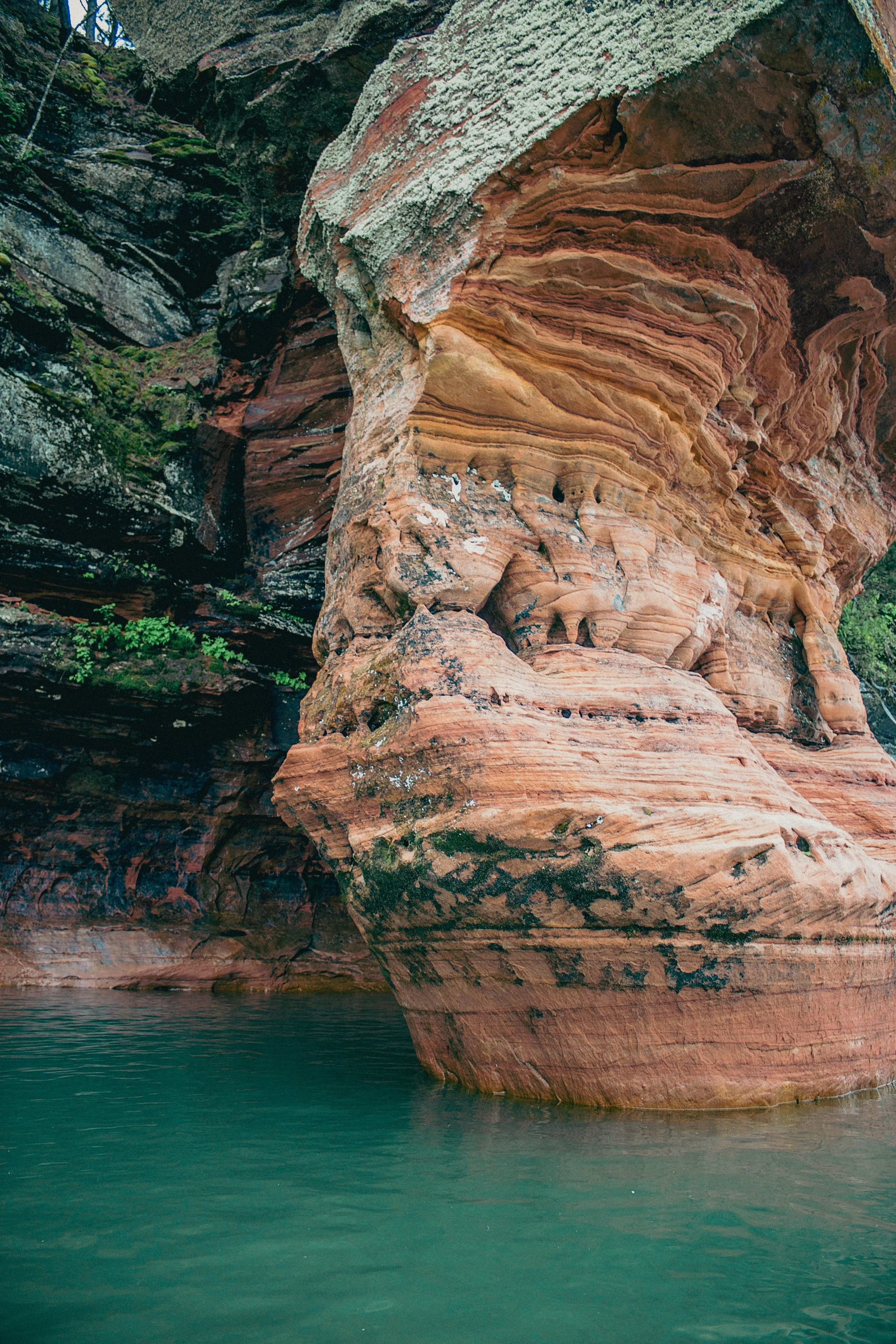 Close-up of a large, reddish-brown rock formation with layered, wavy patterns, reflected in calm green water at its base, with moss and lichen on the surface.