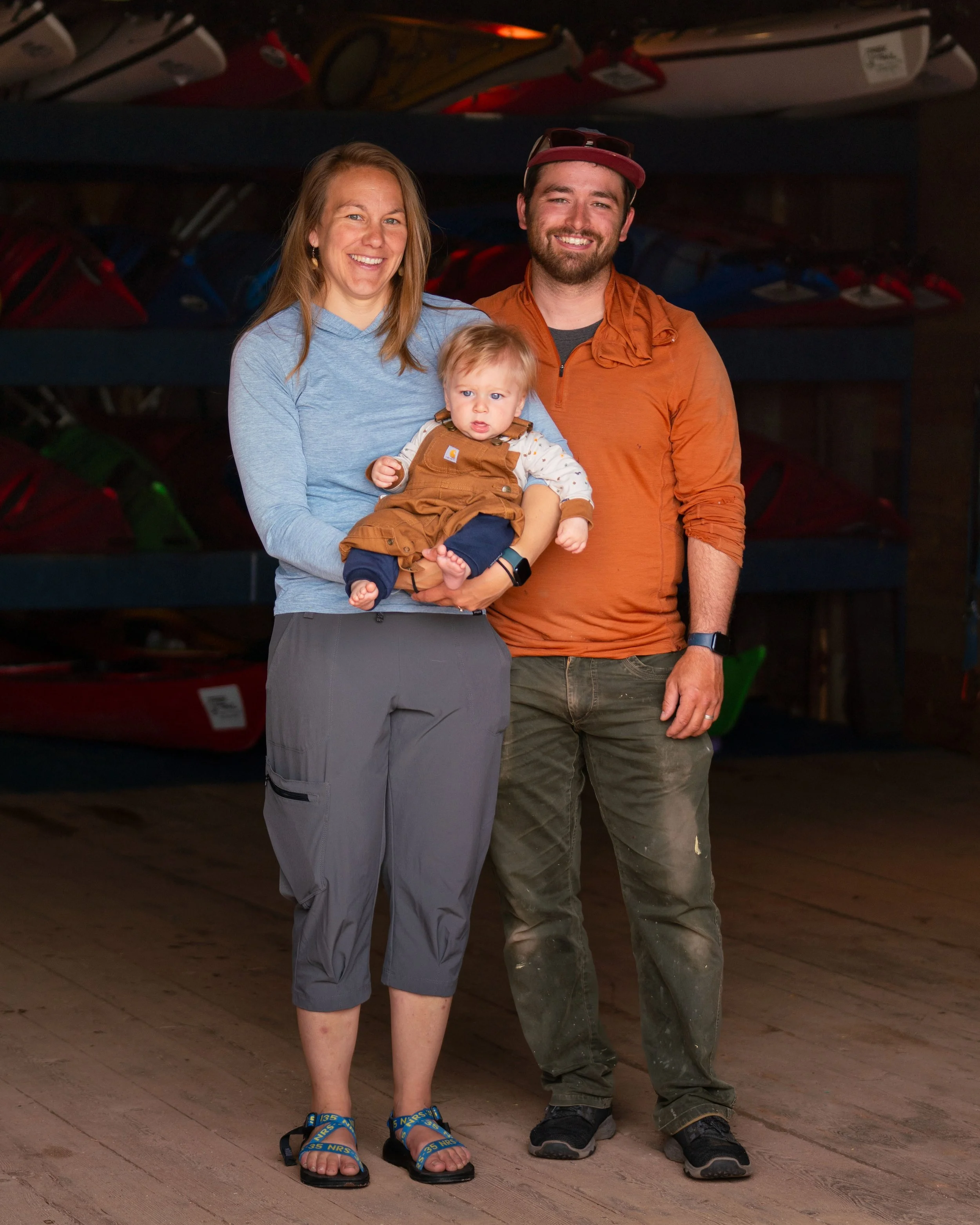A family of three standing inside a store in front of a wall of canoes and kayaks. The woman is holding a young boy, and the man stands next to her. They are smiling at the camera.