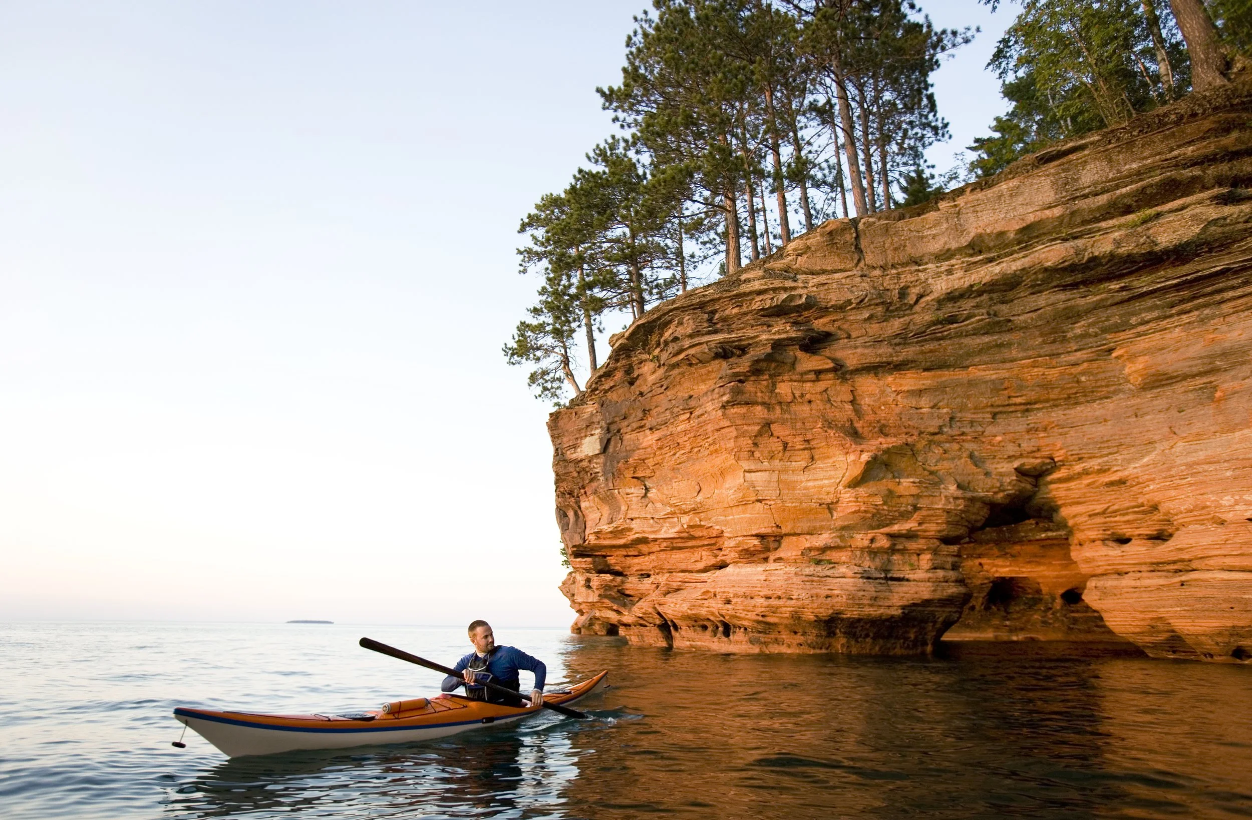 A man kayaking near a large rocky cliff with trees on top, on a calm body of water during sunset or sunrise.