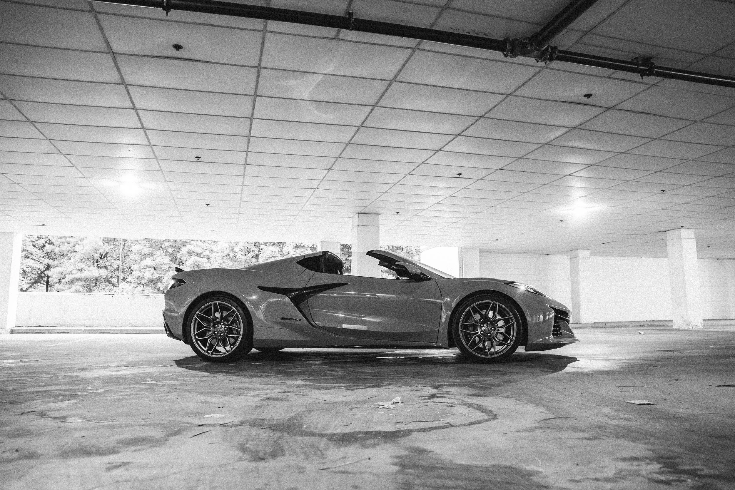 A sleek sports car parked in an empty, covered parking lot with a grid ceiling, concrete pillars, and large windows showing trees outside. The car is low, with a aerodynamic design and shiny surface.