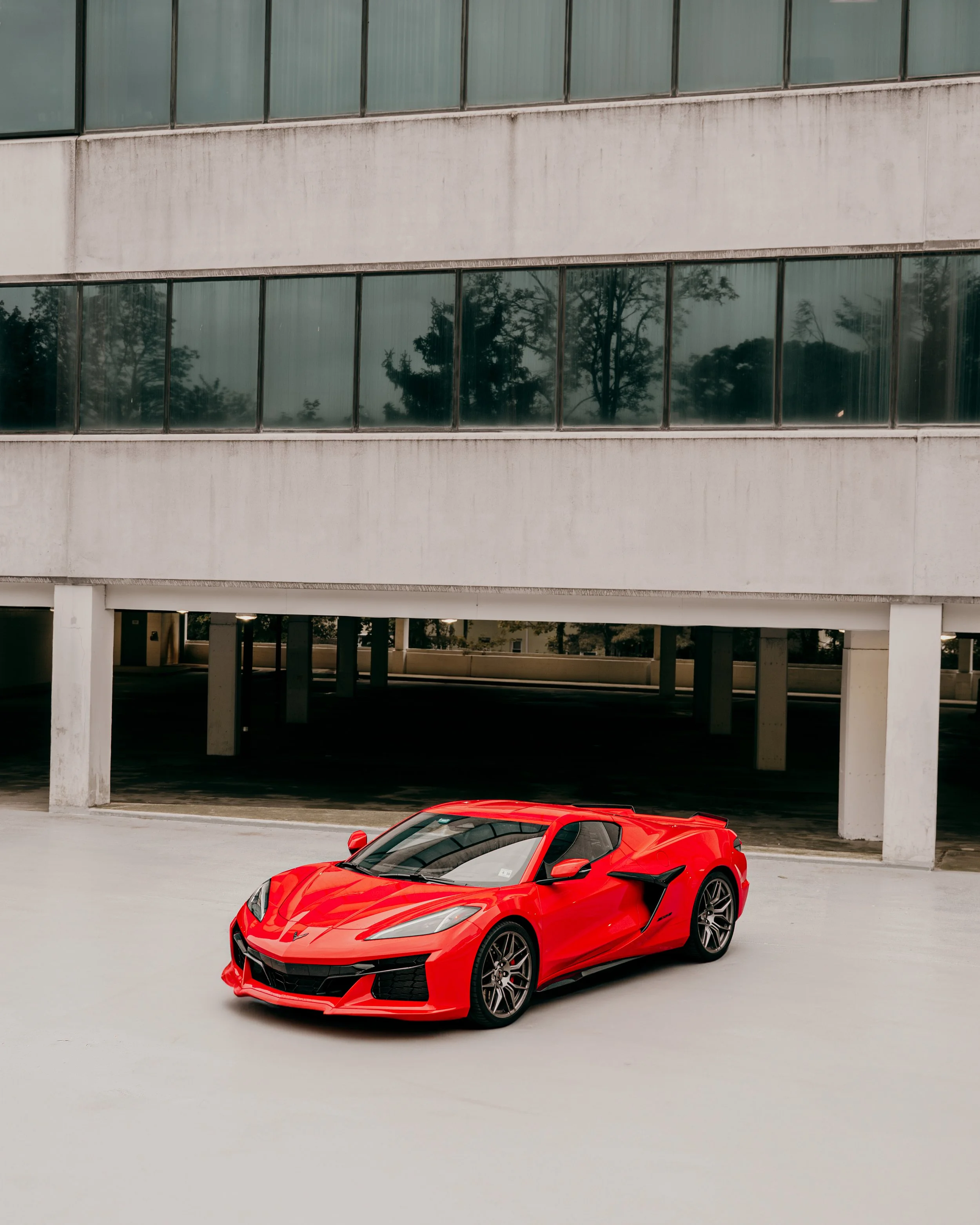 A red sports car parked on a light gray concrete surface with a modern concrete and glass building in the background.