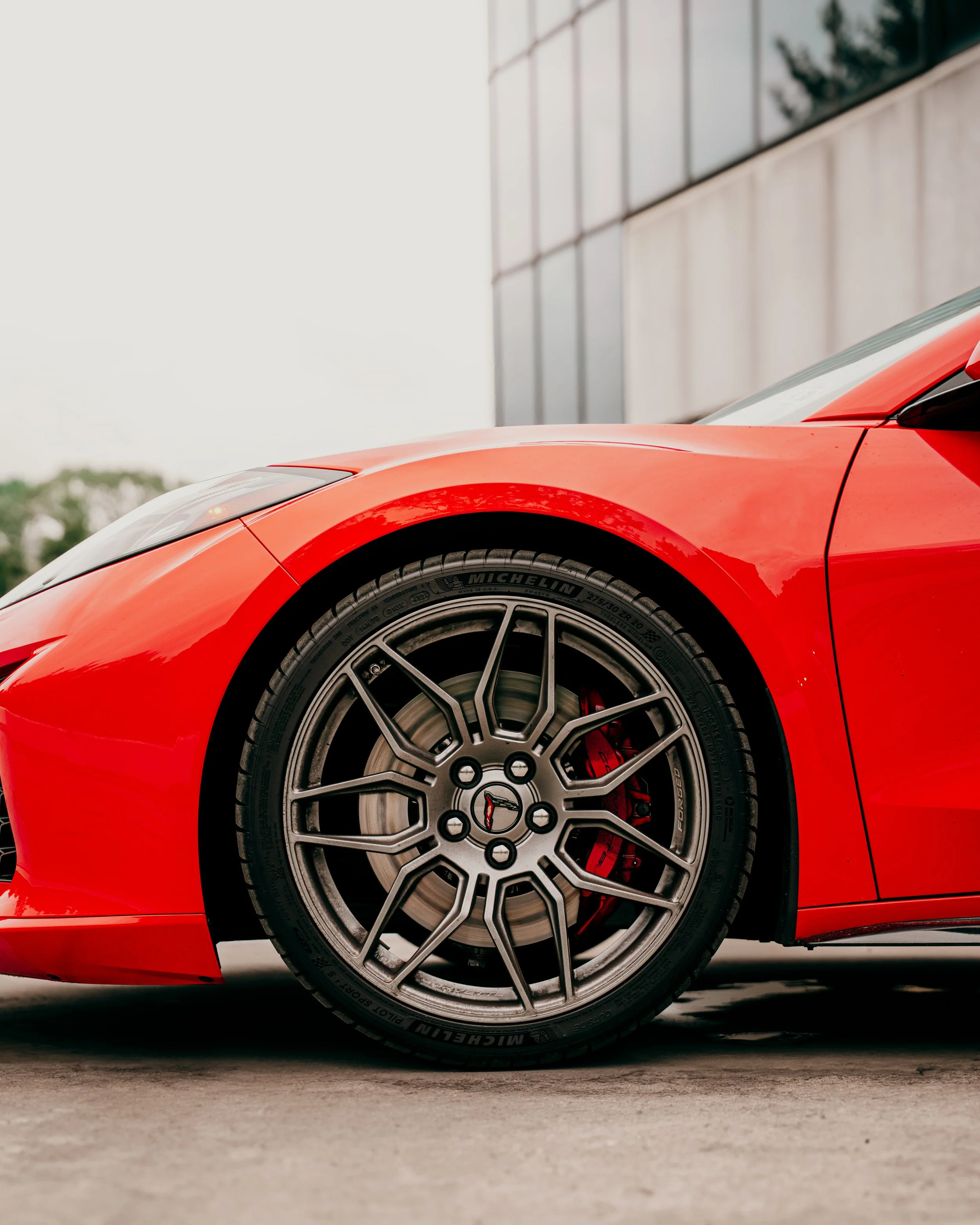Close-up of front part of a red sports car, focusing on the wheel, with a low-profile tire and a black alloy rim, parked on a concrete surface near a building with reflective windows.