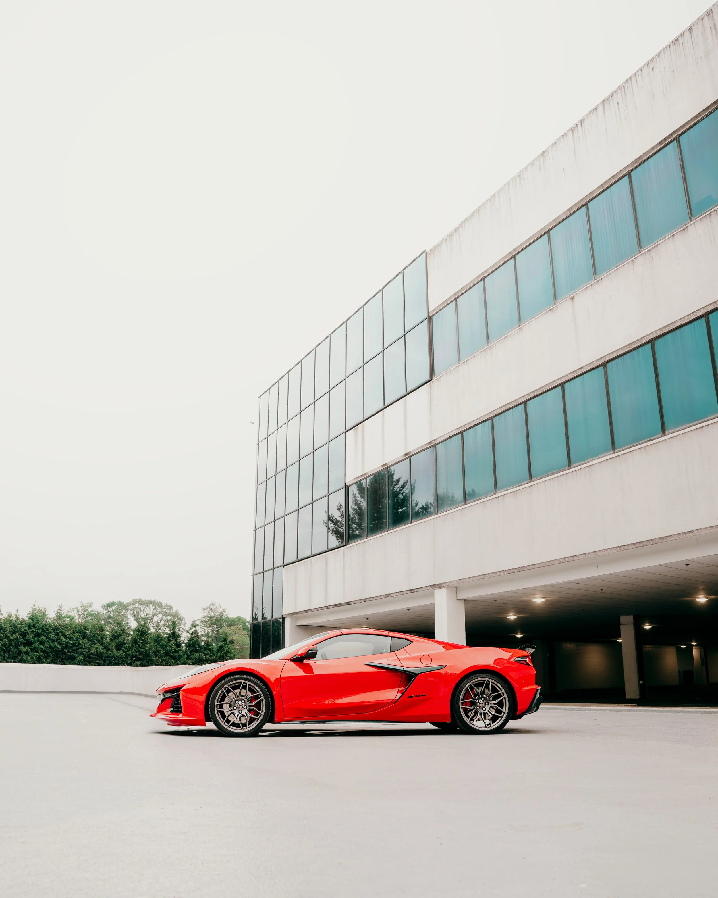 Red sports car parked under a modern building with glass windows and white exterior, with trees in the background.