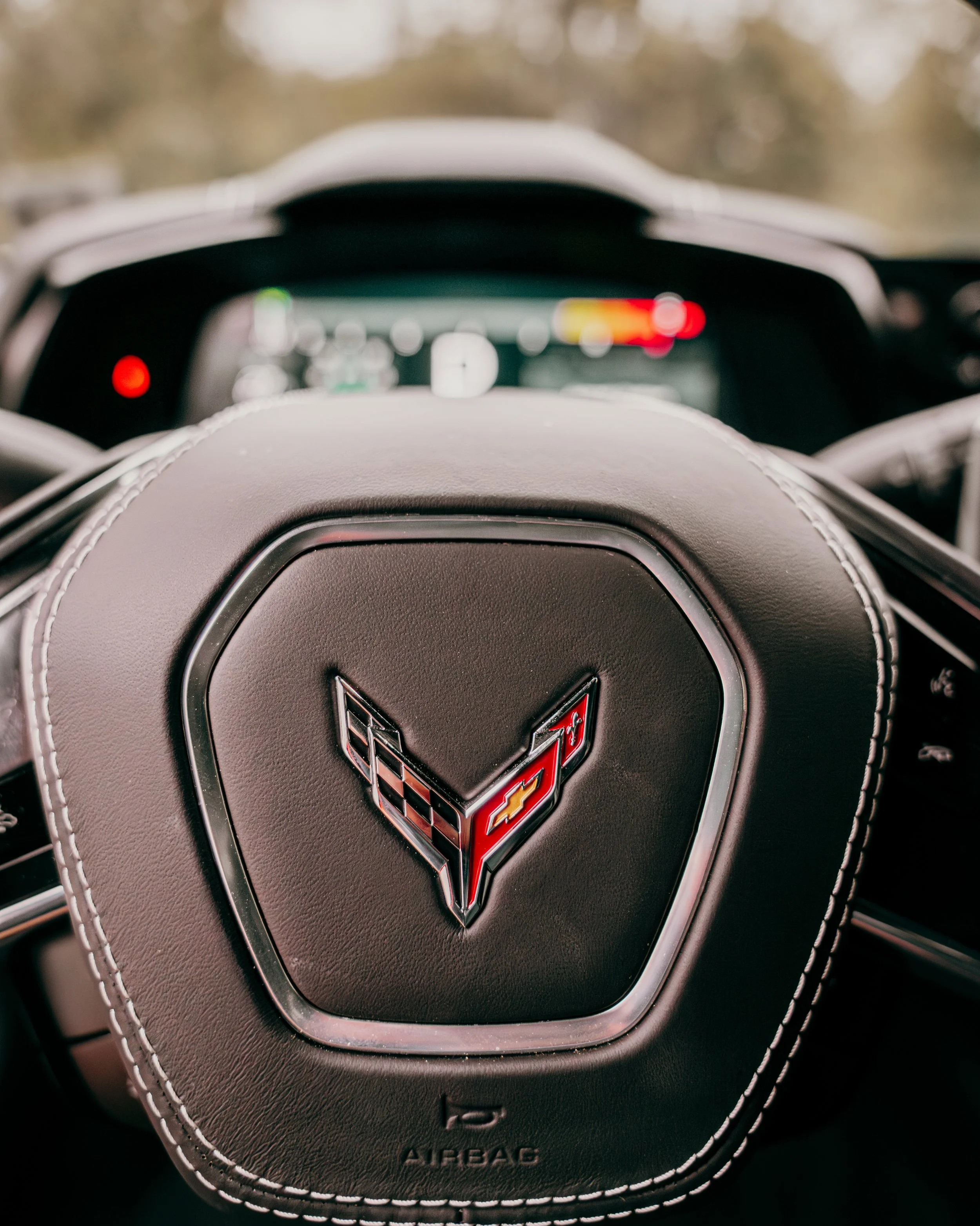 Close-up view of a Chevrolet Corvette steering wheel with a Chevrolet emblem and 'AIRBAG' text, stitched leather design, and a blurred dashboard in the background.