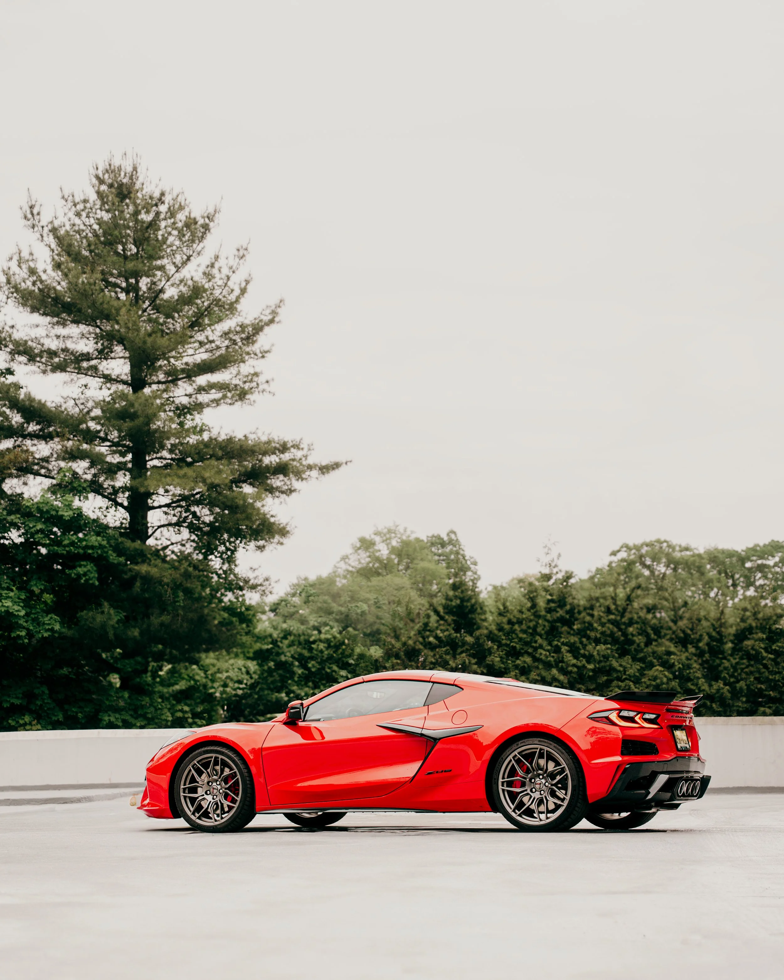 A bright red sports car parked on a white concrete surface with green trees and a cloudy sky in the background.