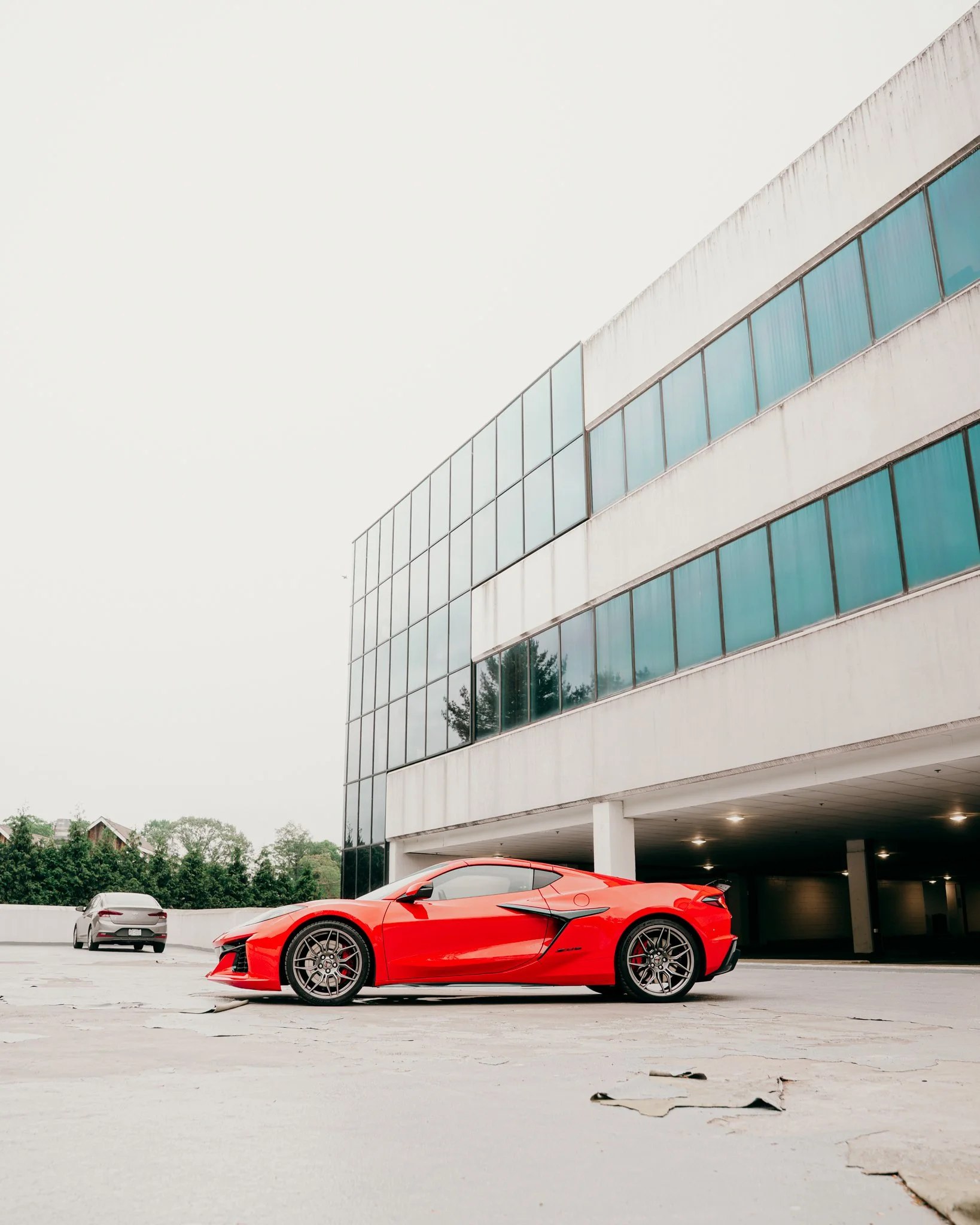 A red sports car parked under a modern building with glass windows, with two other cars in the background, in a spacious parking lot.
