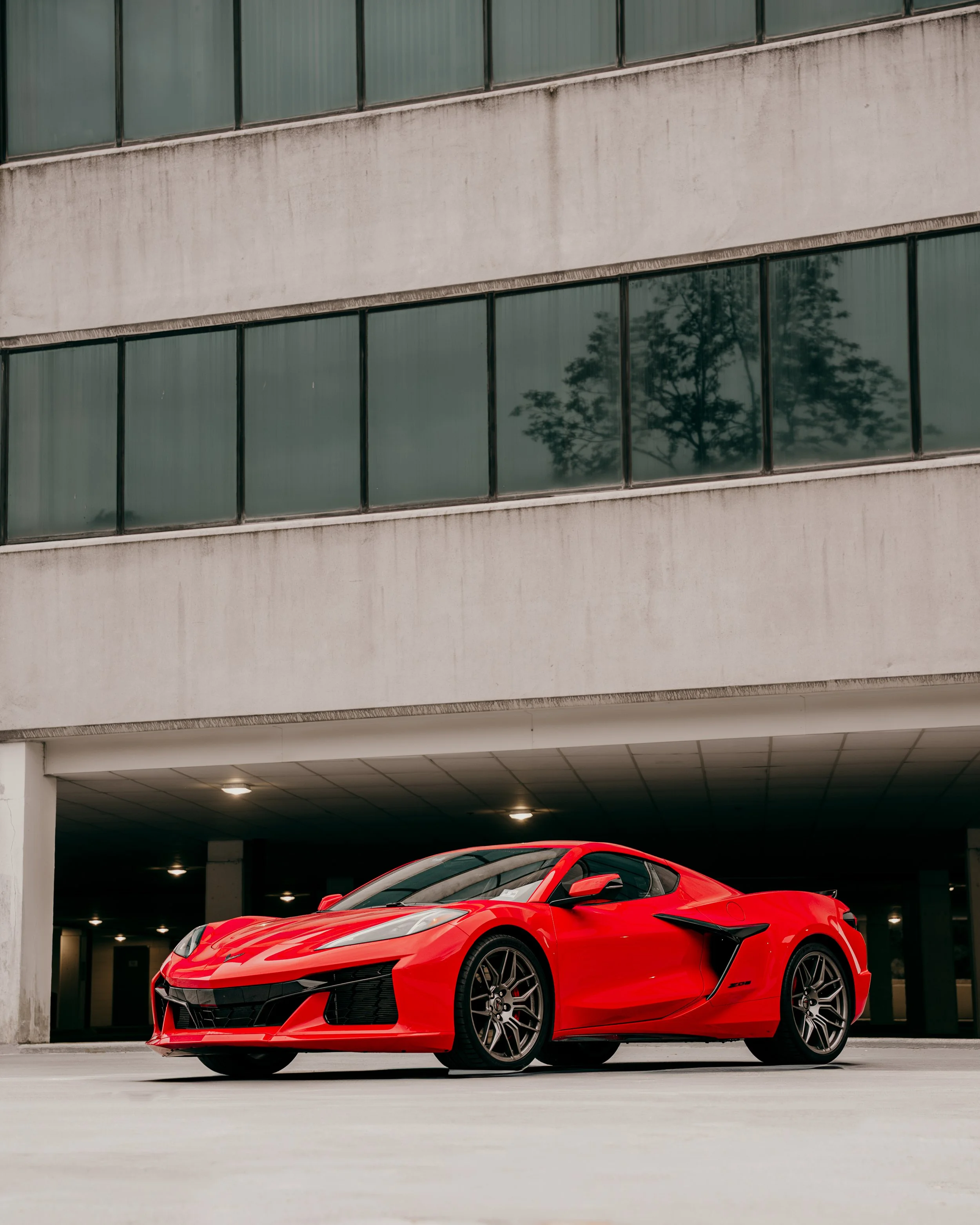 A red sports car parked under a parking garage, with a modern building and trees reflected in the windows above.