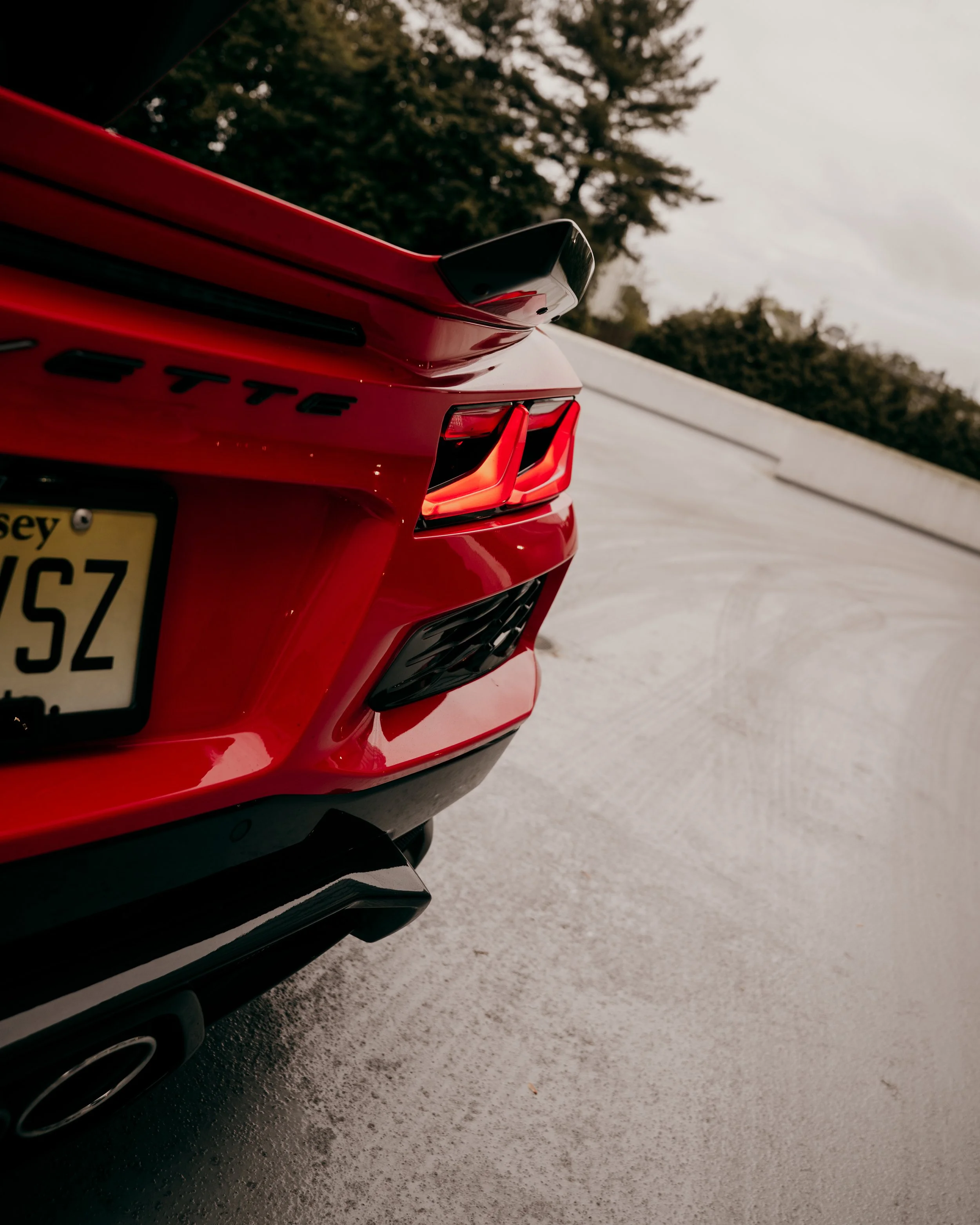 Close-up of a red sports car's rear with a black spoiler, tail light, and part of the exhaust pipe, on a paved road with trees and overcast sky in the background.