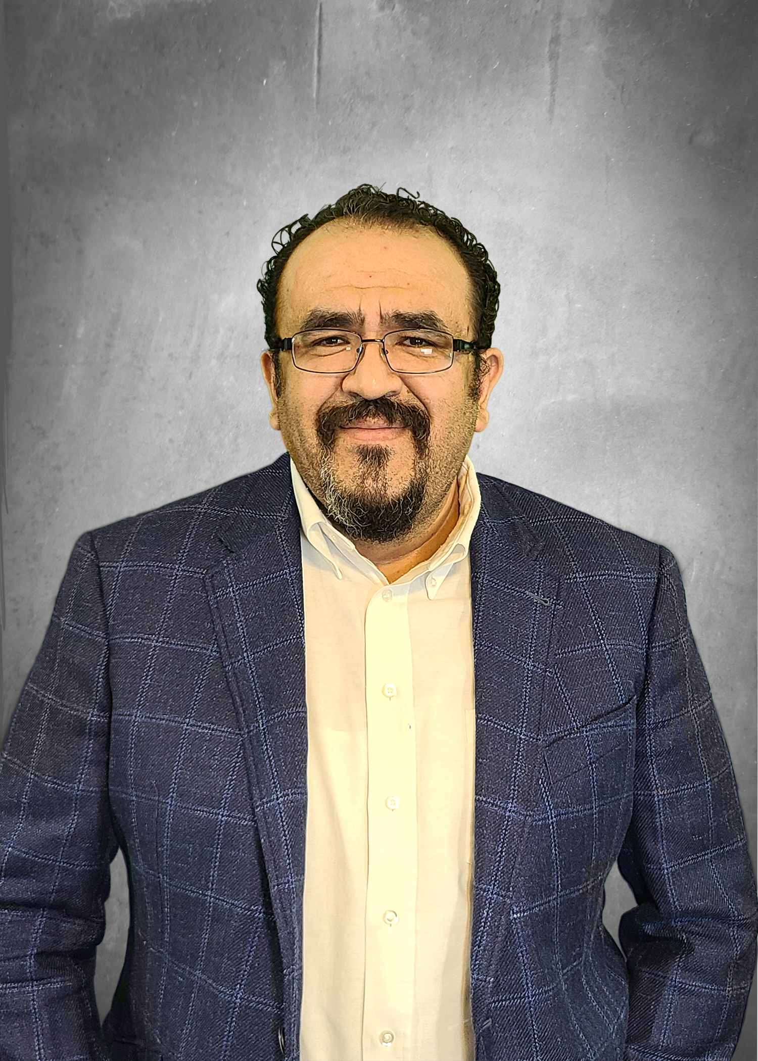 A man with dark curly hair, glasses, and a goatee smiling, wearing a white shirt and a blue checkered blazer, sitting against a plain grey background.