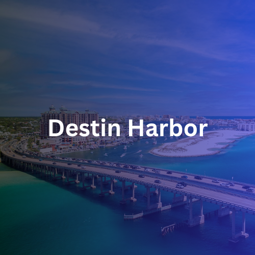 Aerial view of Destin Harbor, Florida, showing a bridge, boats on the water, and buildings along the shoreline under a blue sky.