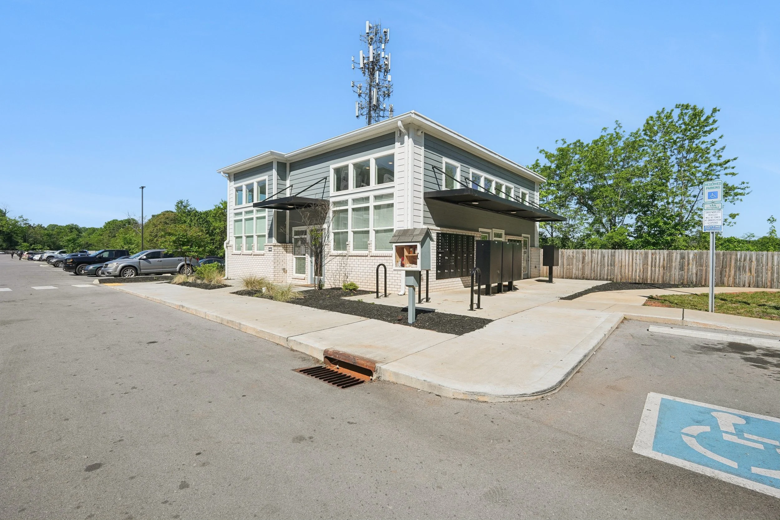 A small building with multiple mailboxes in a parking lot, with a parking space reserved for handicapped nearby and a background of trees and a blue sky.