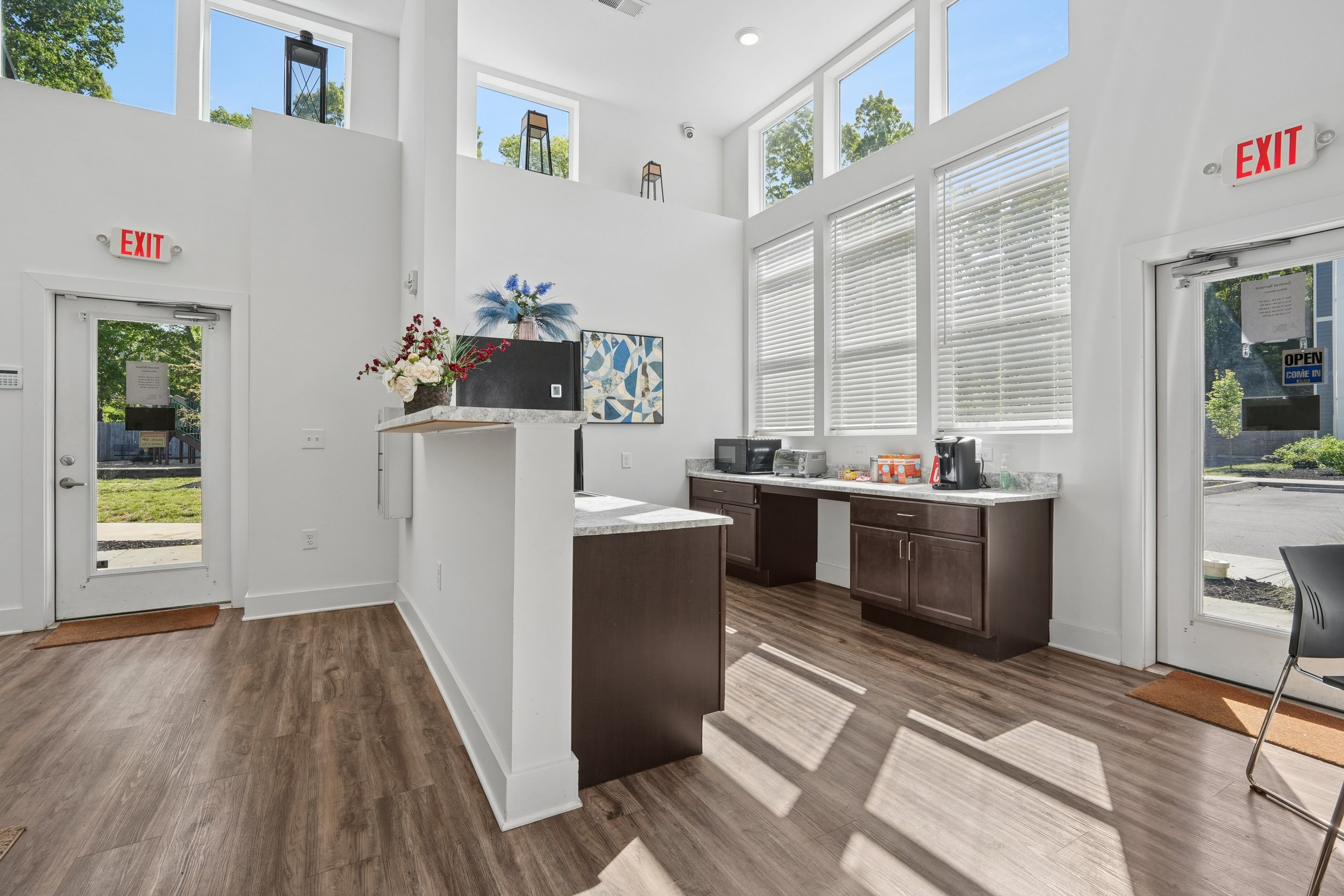 Interior of a modern commercial kitchen with white walls and large windows, brown cabinets, a marble countertop, and a coffee machine on the counter.