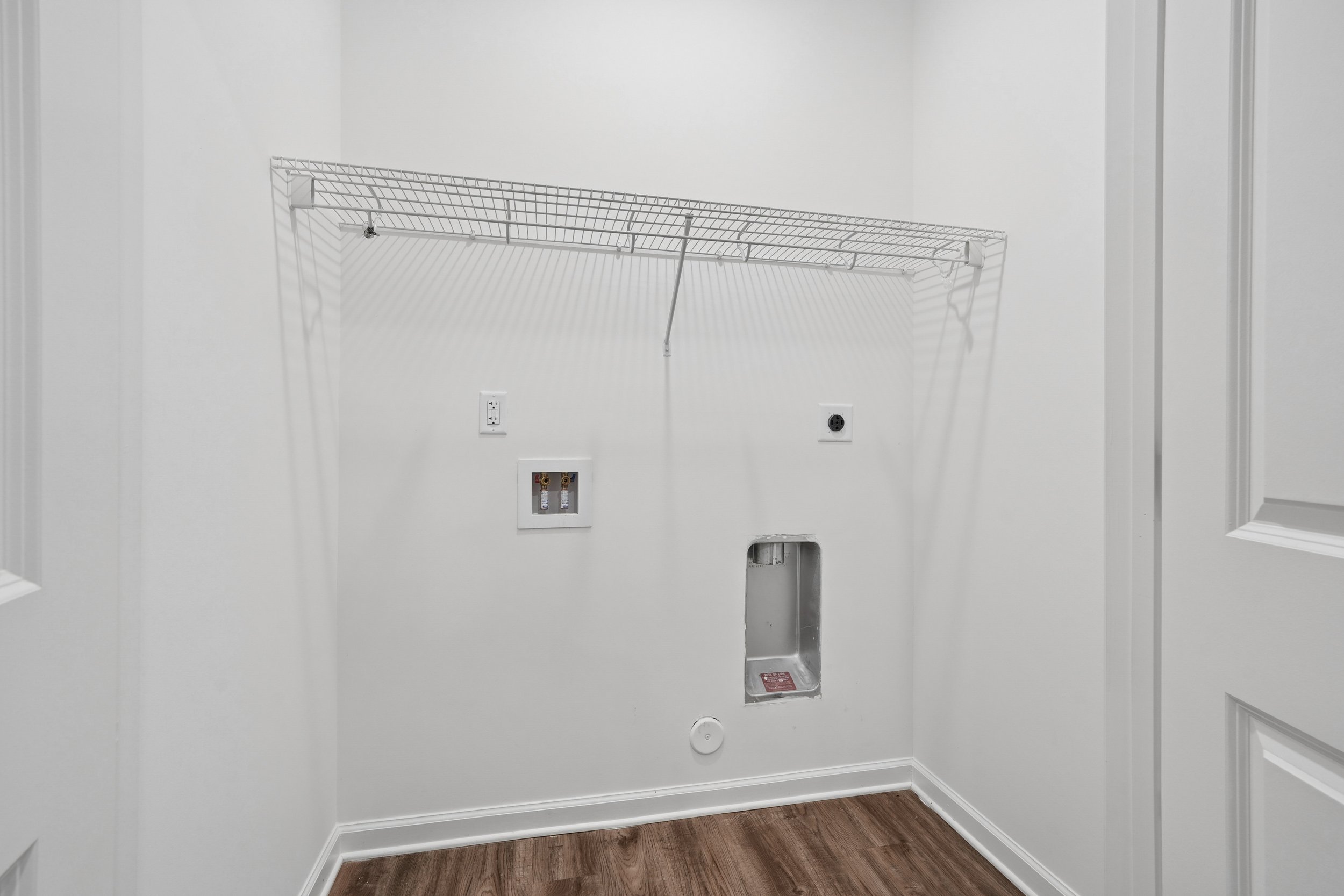 Empty laundry room with a wire shelf, electrical outlets, a vent, and plumbing connections for a washer and dryer, featuring white walls and wood flooring.