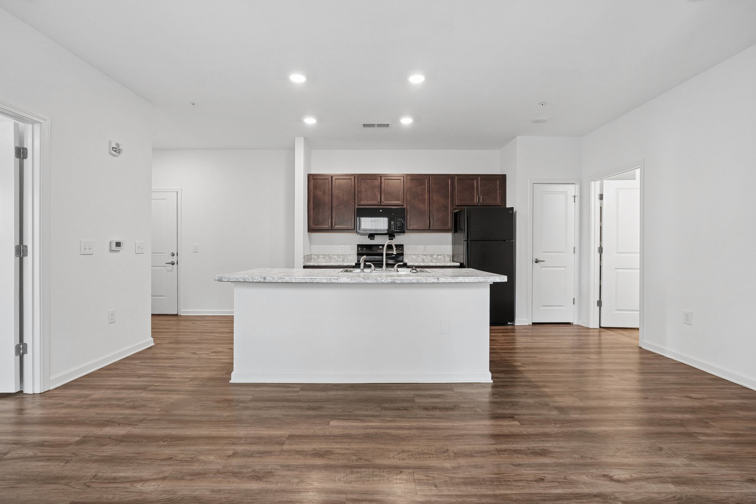Empty modern kitchen with brown cabinets, black appliances, granite countertops, white walls, and wood flooring.