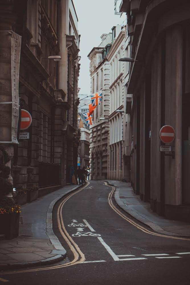 A narrow city street with a bike lane marked on the road, lined with tall historic buildings, British flags hanging between buildings, and a group of people walking in the distance.