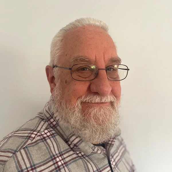 Close-up of an older man with white hair and beard wearing glasses and a plaid shirt, smiling against a plain light-colored background.