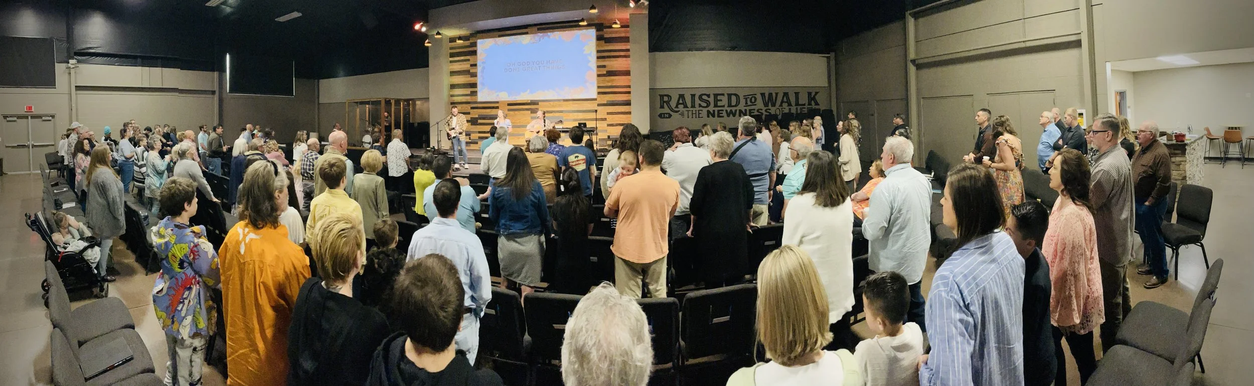 A crowd of people gathered in a church auditorium for a worship service, with a band performing on stage in front of a large screen and a wall with the words 'RAISED TO WALK THE NEWNESS OF LIFE.'