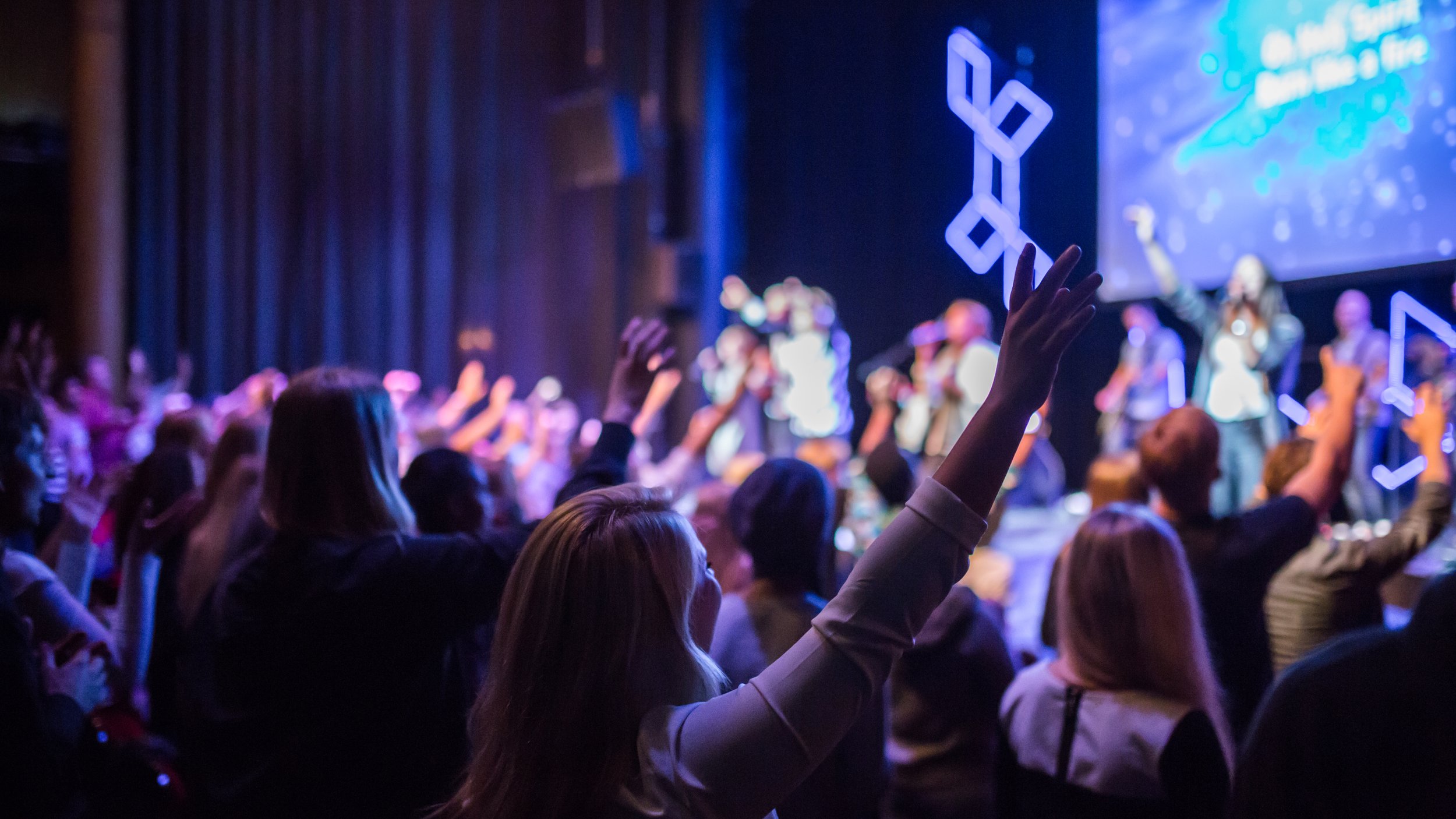 Audience raising their hands during a concert or event on a stage with lighting, with performers in the background and a large screen showing graphics.