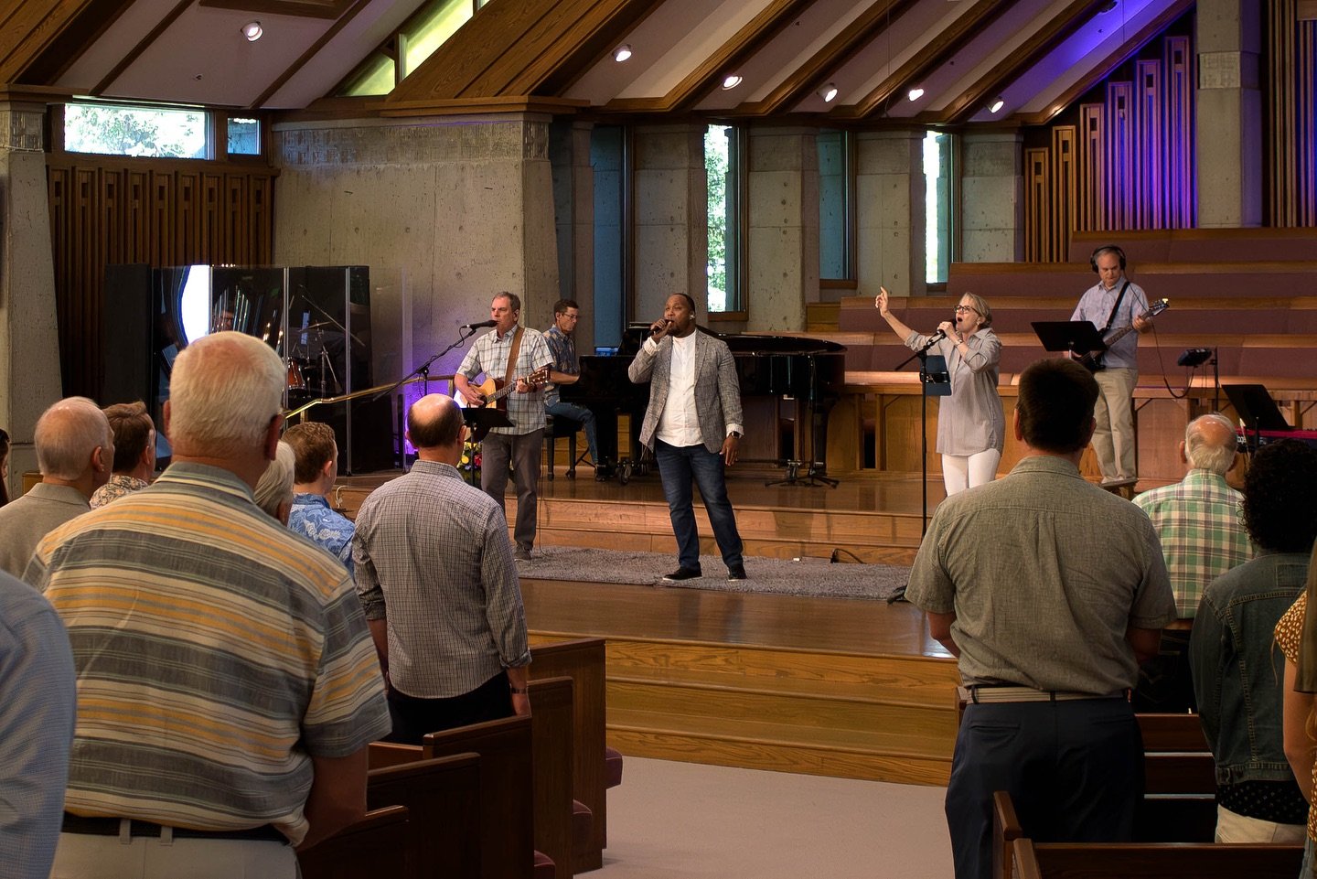 A live band performs on stage in front of an audience inside a church or auditorium, with five band members playing instruments and singing, and people standing and watching attentively.