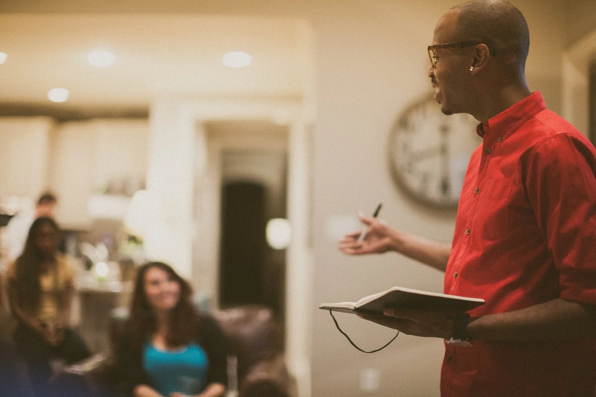 A person in a red shirt and glasses speaking to a group in a living room setting.