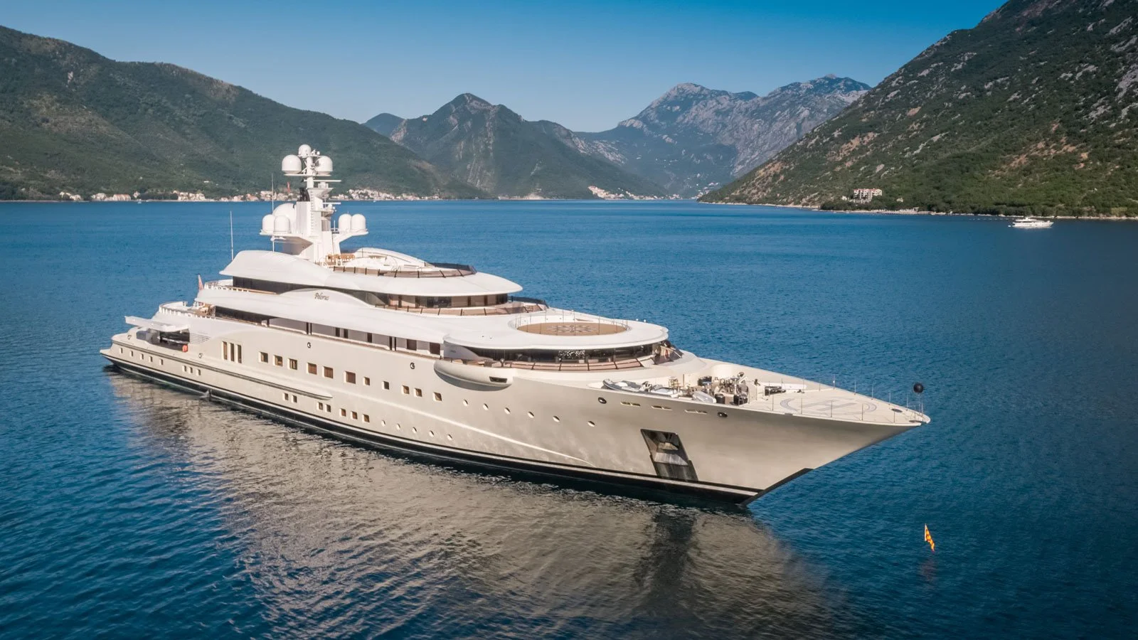 A large white yacht sailing on a calm body of water surrounded by green mountains under a clear blue sky.