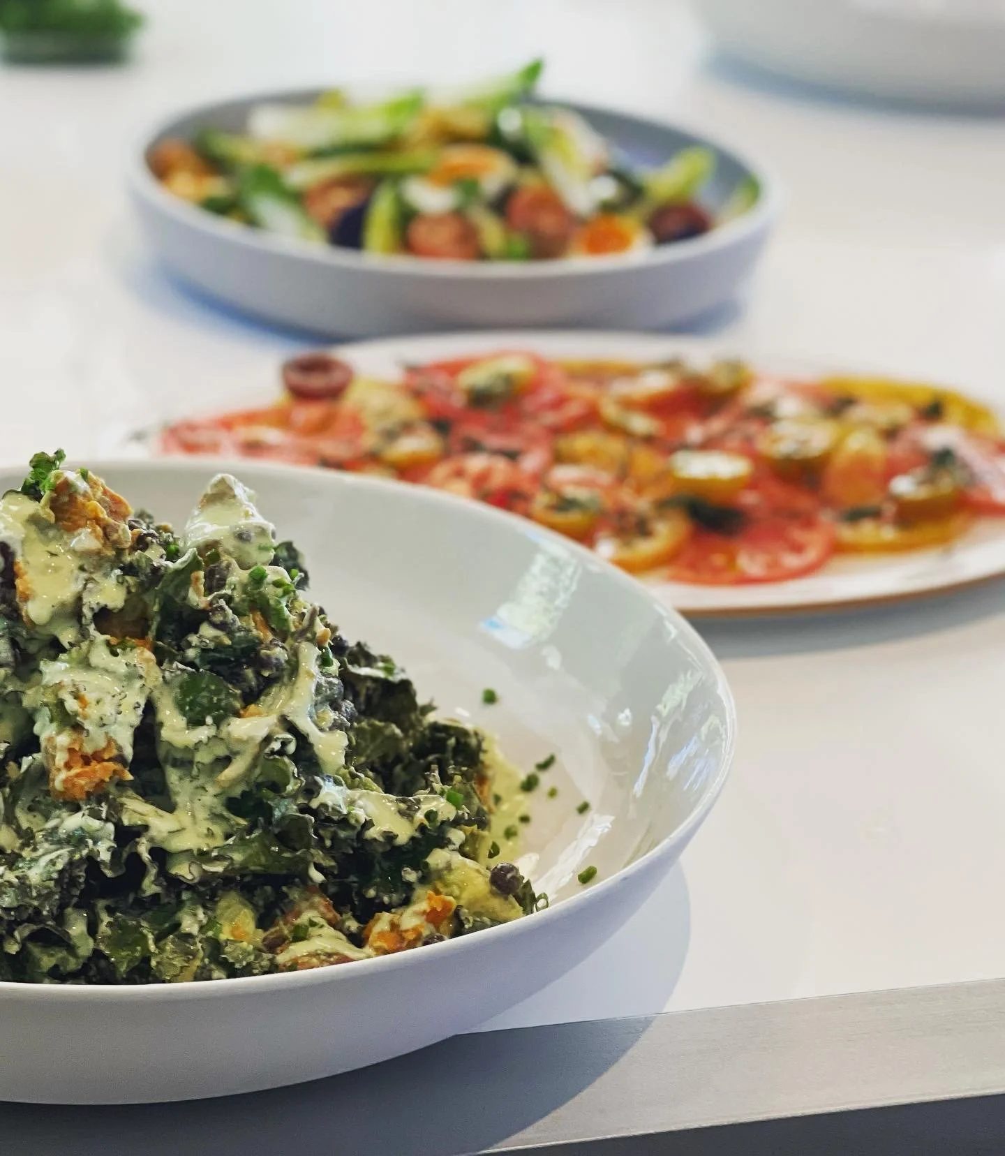 Close-up of a bowl of creamy kale salad with nuts and cheese in the foreground, with a plate of sliced tomatoes and a salad bowl in the background.