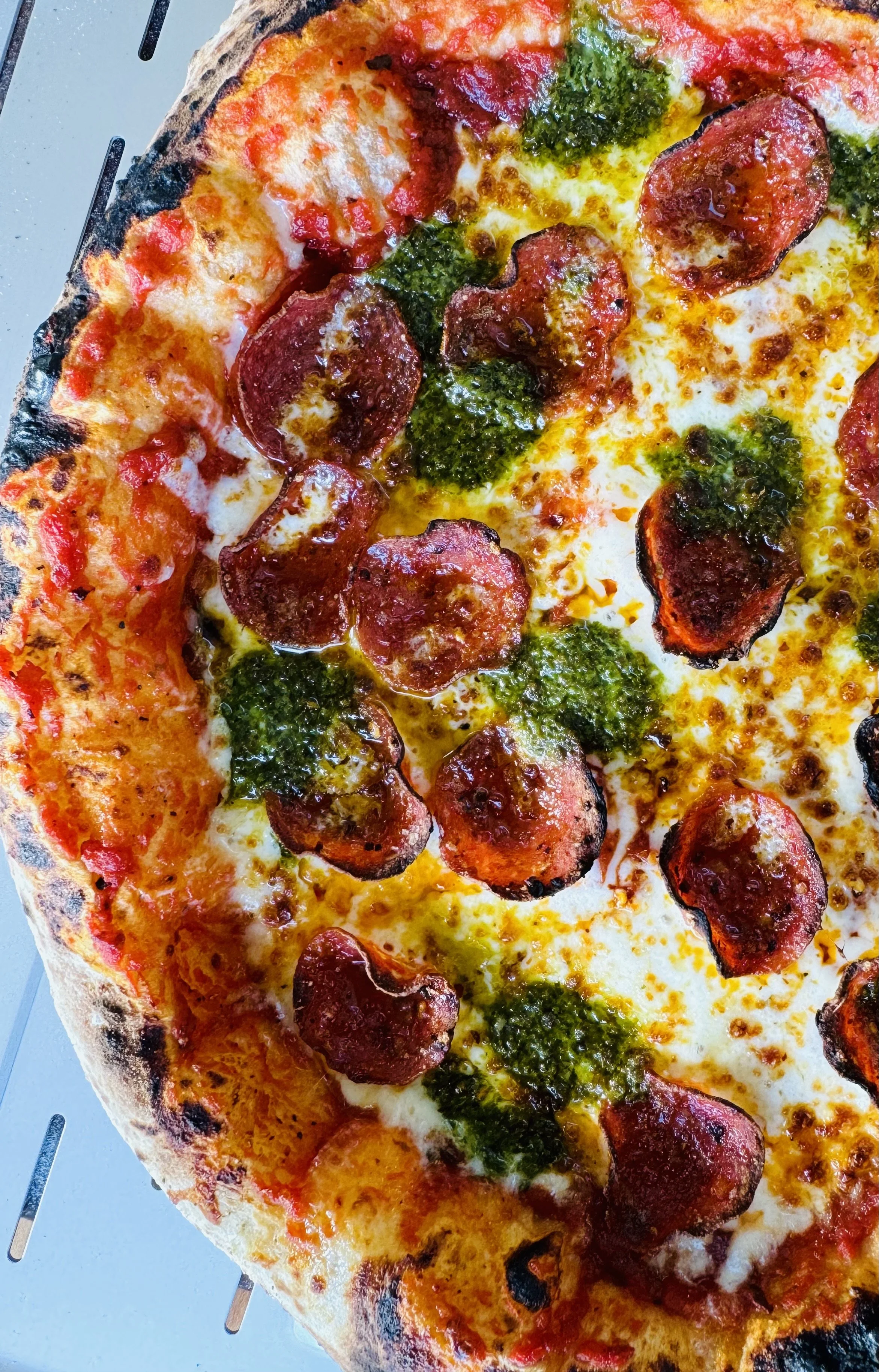 Close-up of a half pizza with pepperoni slices, green pesto, and melted cheese on a metal tray.