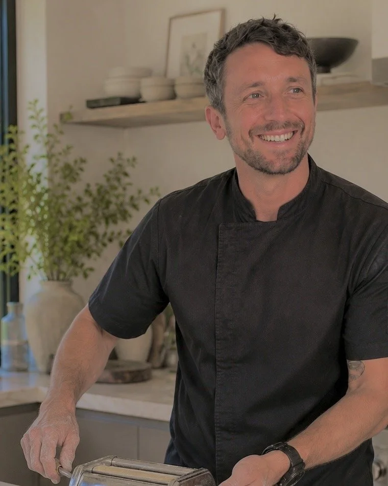 A smiling male chef in a black uniform preparing food in a modern kitchen with plants and decorative items in the background.