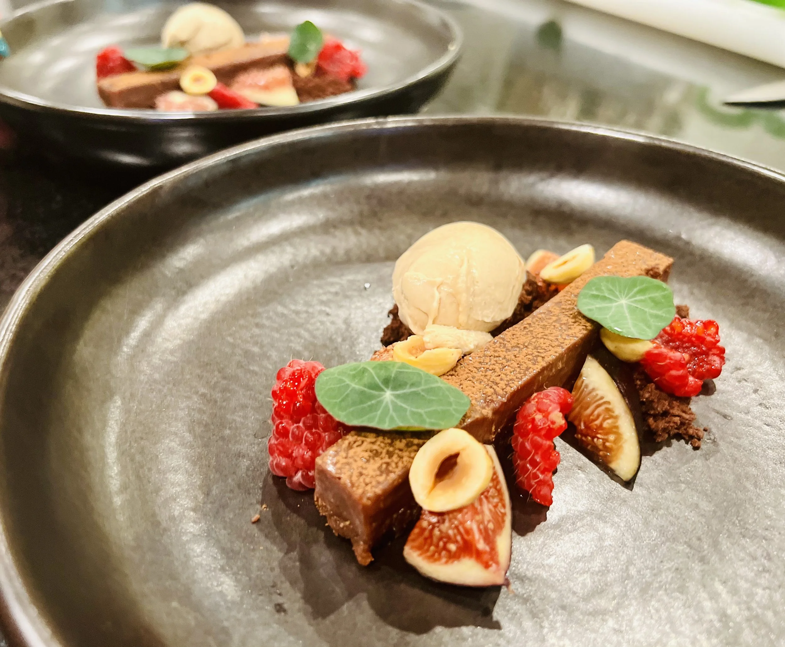 Close-up of plated gourmet dessert with chocolate, fruits, nuts, and edible flowers on black and gray dishes.