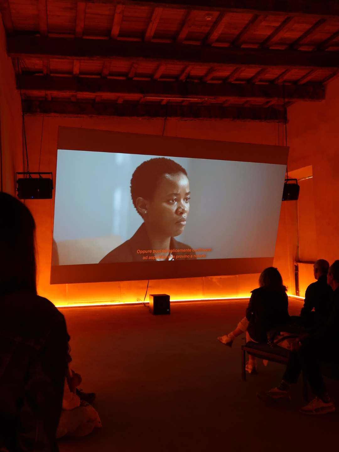 Interno di una sala con schermo che mostra un film, alcune persone sedute e in piedi guardano lo schermo, ambiente con illuminazione calda e soffitto di legno.