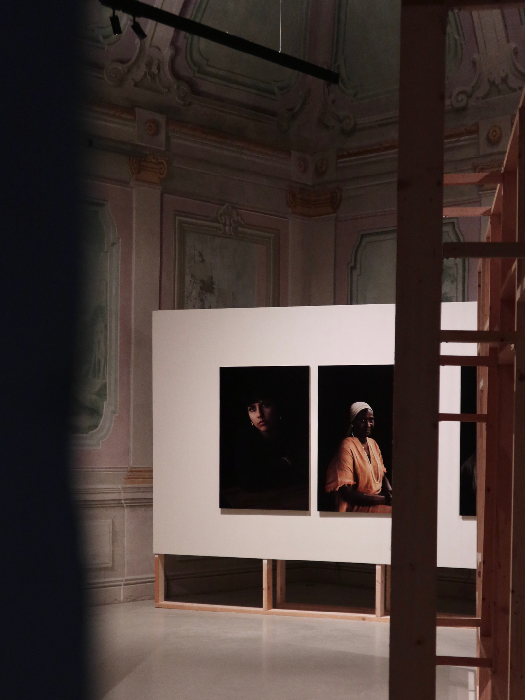 Due fotografie di ritratti di persone su sfondo scuro, esposte su una parete di un museo o galleria, con scaffali di legno visibili sul lato destro e un soffitto decorato in stucco.
