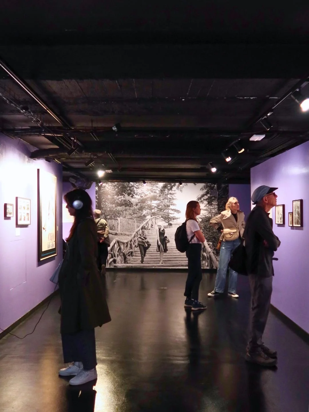 Visitatori in un museo di arte moderna con pareti viola, osservano varie opere e una grande foto in bianco e nero di persone che attraversano un ponte in un ambiente naturale.
