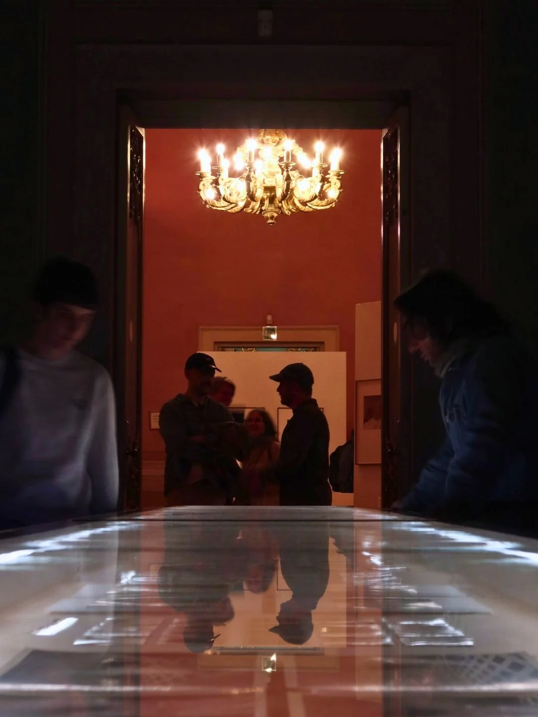 Interno di un museo con un lampadario di cristallo e persone che osservano una mostra, riflessi sul tavolo