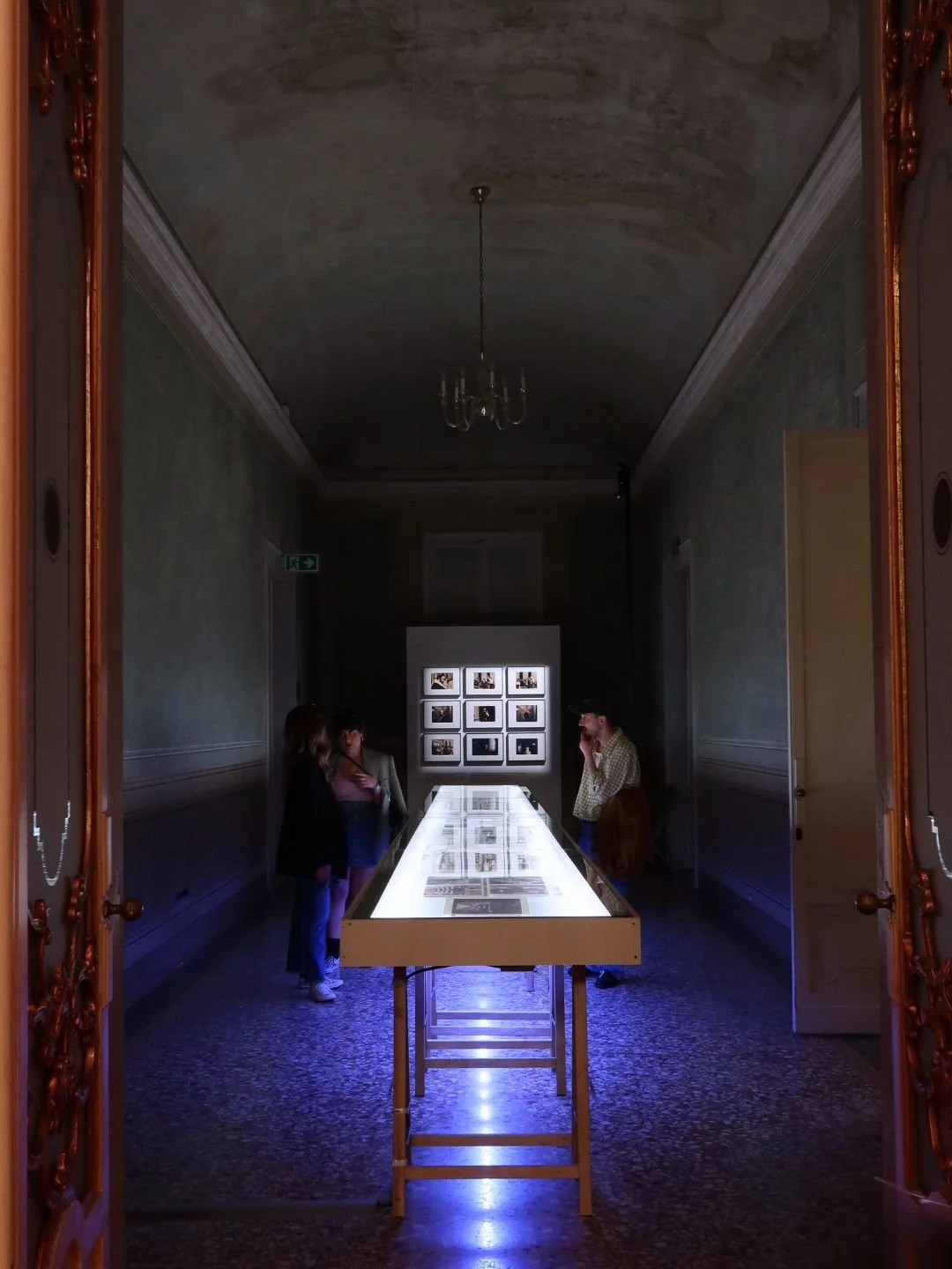 Una stanza d'epoca con pareti color verde chiaro, soffitto curvo e pavimento a mosaico. Al centro, un tavolo di vetro illuminato mostra fotografie o documenti. Sullo sfondo, un pannello con più foto incorniciate e tre persone che osservano le immagin