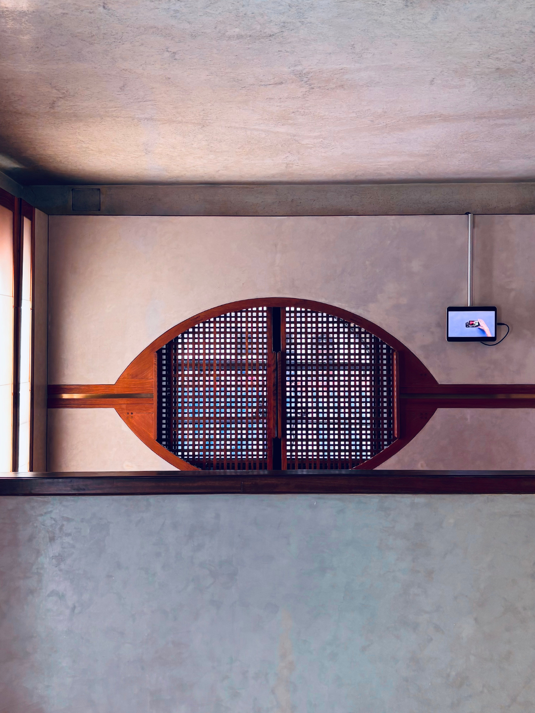 Parete con finestra di forma ovale in legno e persiana a griglia, e un monitor digitale a parete con cavo.